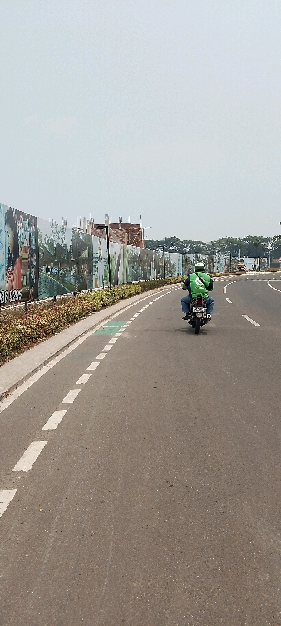 The image shows a motorcyclist wearing a green jacket with the "GOJEK" logo, riding on a road in Gunung Putri, Indonesia. The road has white dashed lines and a green painted lane marking. To the left of the road, there is a decorative fence with large posters, and beyond that, construction is underway with visible building materials and scaffolding. The sky is overcast. The scene captures a typical street view in the suburban area of Gunung Putri, indicating commercial or residential development activities.