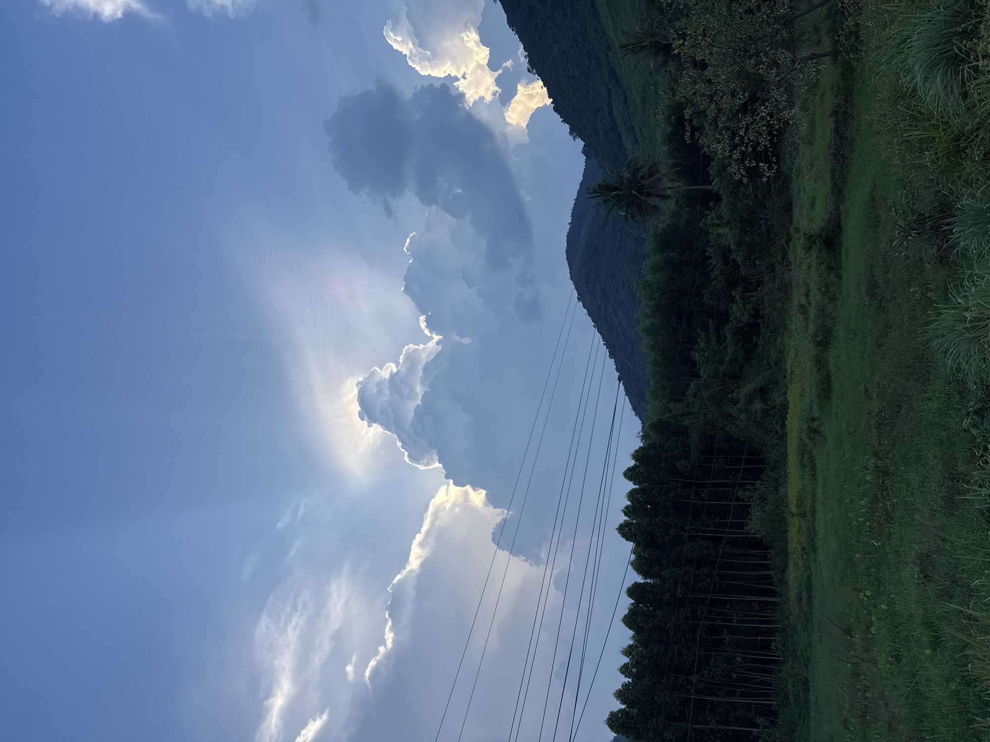 The image shows a landscape in Tây Ninh Hòa, Vietnam. A mountain with dense green vegetation occupies the right side of the frame. Power lines are visible in the middle ground, running diagonally across the image. The left side features a blue sky with large, bright clouds partially obscuring the sun, which is creating rays of light. There are no people visible in the scene.