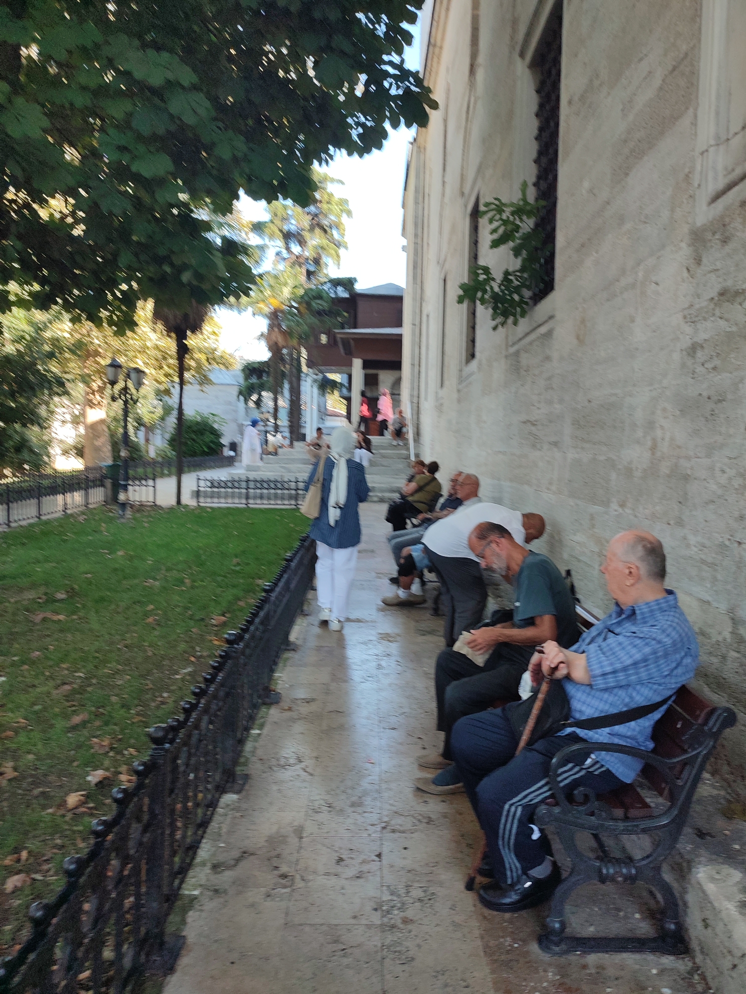 In Üsküdar, Turkey, a group of men are seated on a bench next to a stone building. A woman in a blue jacket and white pants, wearing a headscarf, walks away from the camera. Other people are visible in the background, some walking up stairs. Trees and a black metal fence line a grassy area to the left of the walkway. The ground is paved with stone and appears wet. The building has a textured stone facade with barred windows.