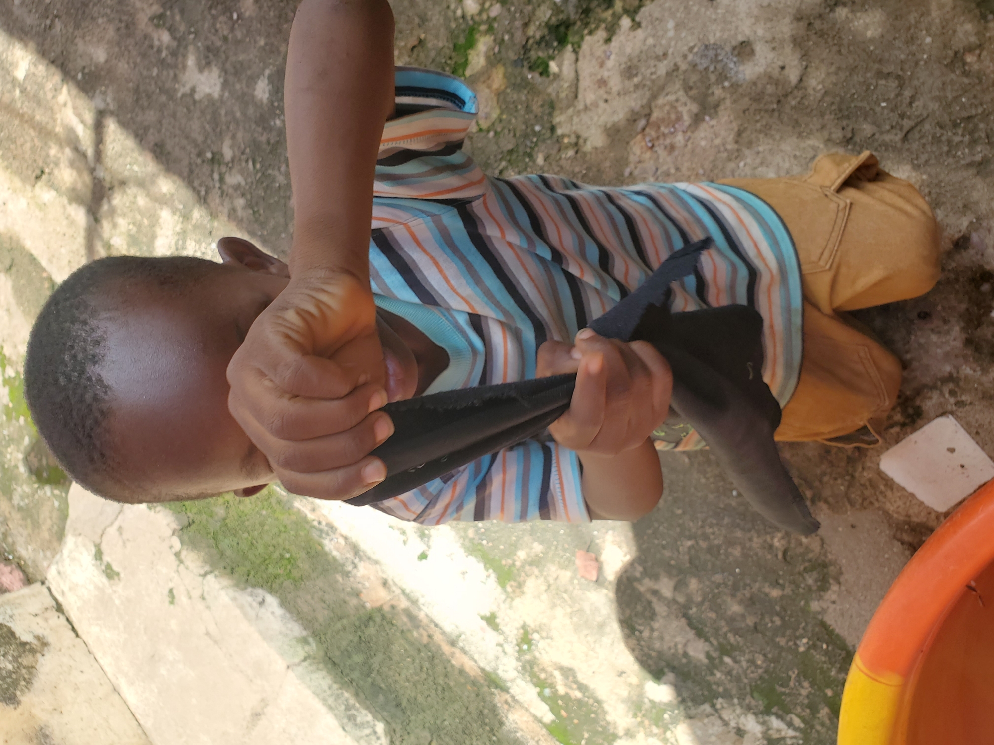 A young boy, estimated to be between 4-6 years old, kneels on a rough, paved outdoor surface, likely in Jos, Nigeria. He is wearing a horizontally striped t-shirt with colors including blue, orange, black, and white, and light brown cargo shorts. The boy is holding a black piece of fabric, possibly a sock or cloth, in both hands, bringing it towards his face and obscuring his eyes. His hands appear to be squeezing or manipulating the fabric. The ground is a mix of concrete and dirt, with some patches of moss or algae visible on the concrete sections. In the lower left of the frame, a portion of a bright orange and yellow plastic basin is visible. The background shows more of the weathered, textured ground and hints of a wall or structure. The overall lighting suggests it is daytime.