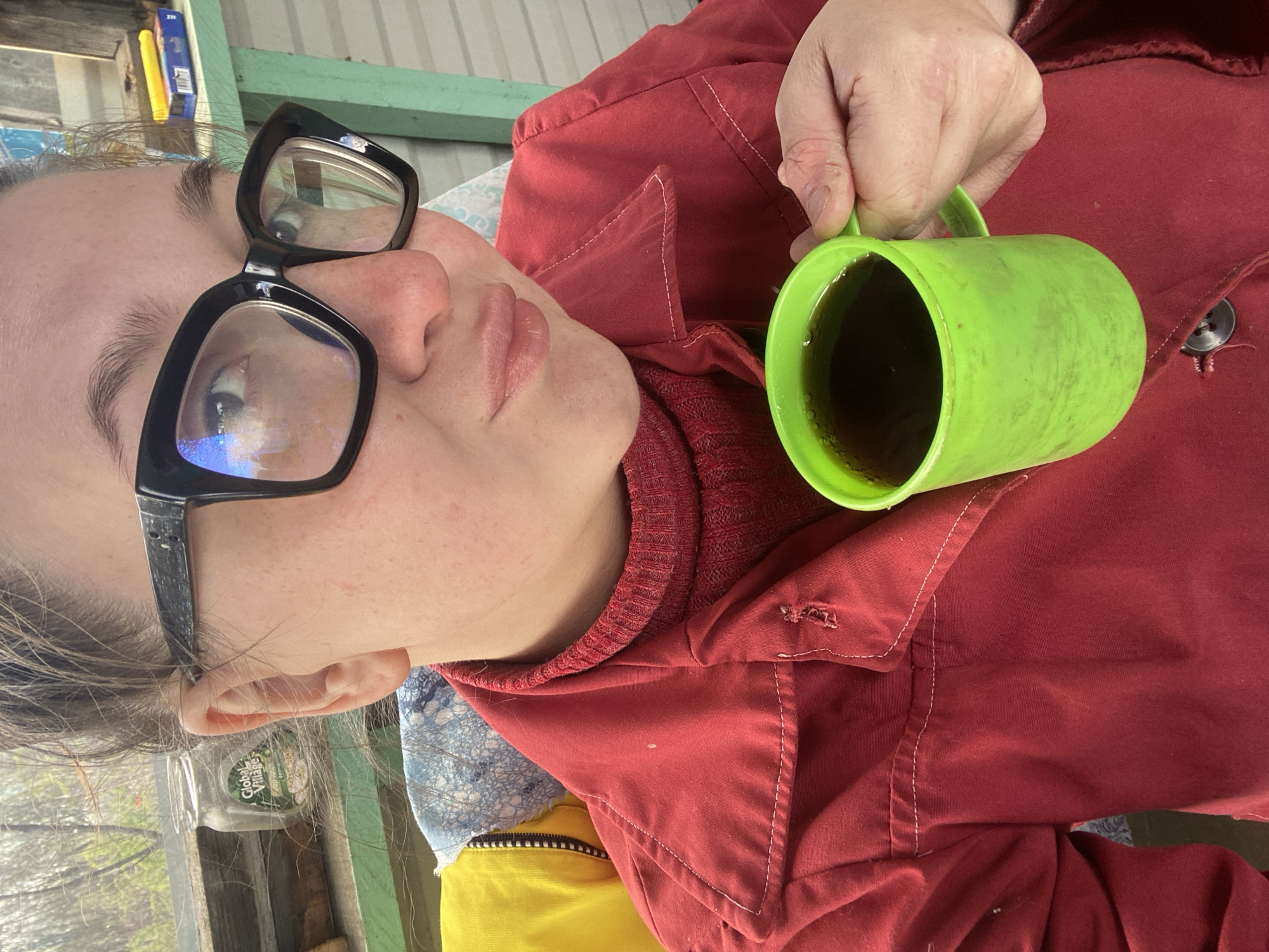 A person wearing black-framed glasses and a red collared jacket, possibly a shirt or light coat, is holding a green plastic mug filled with a dark liquid. They are indoors and the image is taken at an oblique angle, resulting in a slightly distorted perspective. The person's face is visible, and they appear to be looking directly at the camera. Patches of yellow and blue fabric are visible near the collar of the red jacket. The background is indistinct. The location and the city in Chaikovsky, Russia are indeterminable.