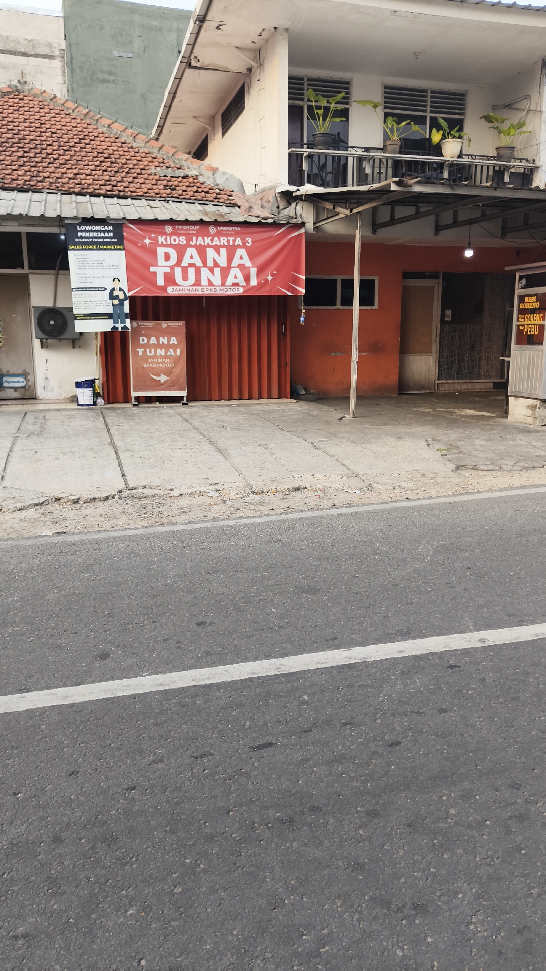 The image depicts a storefront in South Jakarta, Indonesia, featuring a bright red banner with white text that reads "Kios Jakarta 3 DANA TUNAI JAMINAN BPKB MOTOR". To the left of the banner is a smaller sign that says "LOWONGAN PEKERJAAN FIFGROUP PASAR MINGGU SALES FORCE / MARKETING". A brown standing sign in front of the red shuttered store also displays "DANA TUNAI JAMINAN BPKB MOTOR". An air conditioning unit is visible on the left wall. A potted plant sits on a balcony above the storefront. In the foreground, a grey asphalt road with a white line runs horizontally. The building appears to be a commercial establishment on a street.