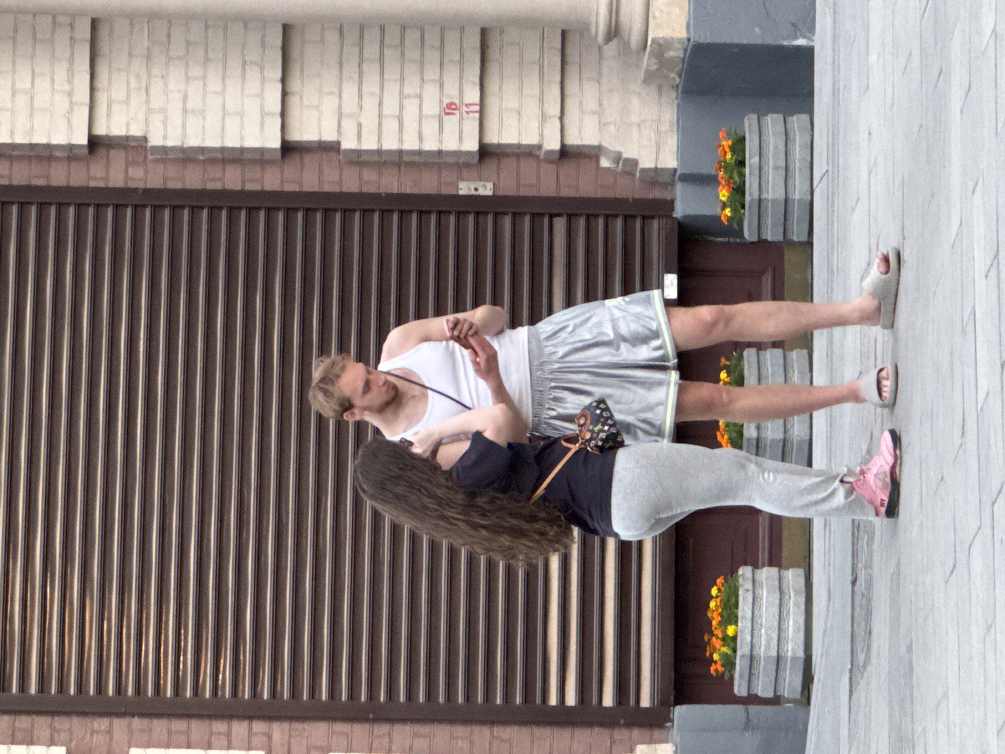 Two individuals, a male and a female, stand outside a building in what appears to be Moscow, Russia. The male is fair-skinned with blond hair and wears a white tank top and silver athletic shorts. He is barefoot, wearing flip-flops. The female has long, dark hair and wears a dark t-shirt, light gray sweatpants, and pink sneakers. She is wearing a small black purse. The couple is positioned in front of a building with a brown slatted metal security gate and a cream-colored brick facade above. There are potted plants with orange and yellow flowers flanking a brown door. The surrounding area is a gray sidewalk. The image is taken from a slightly elevated perspective.