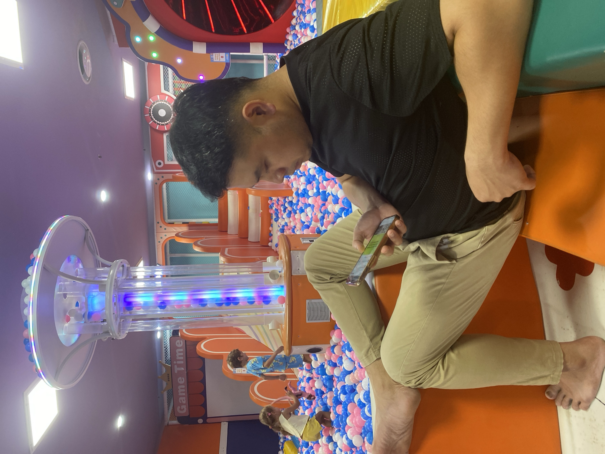 In an indoor ball pit area with colorful walls and lighting, a man is seated, looking at his phone. He is wearing a black t-shirt and beige pants, with one bare foot visible. Two children are playing in the ball pit behind him. The background includes a brightly lit cylindrical structure with colorful balls inside, and other playground equipment. The context suggests this is a children's entertainment venue, potentially in Phu Thuy, Vietnam.
