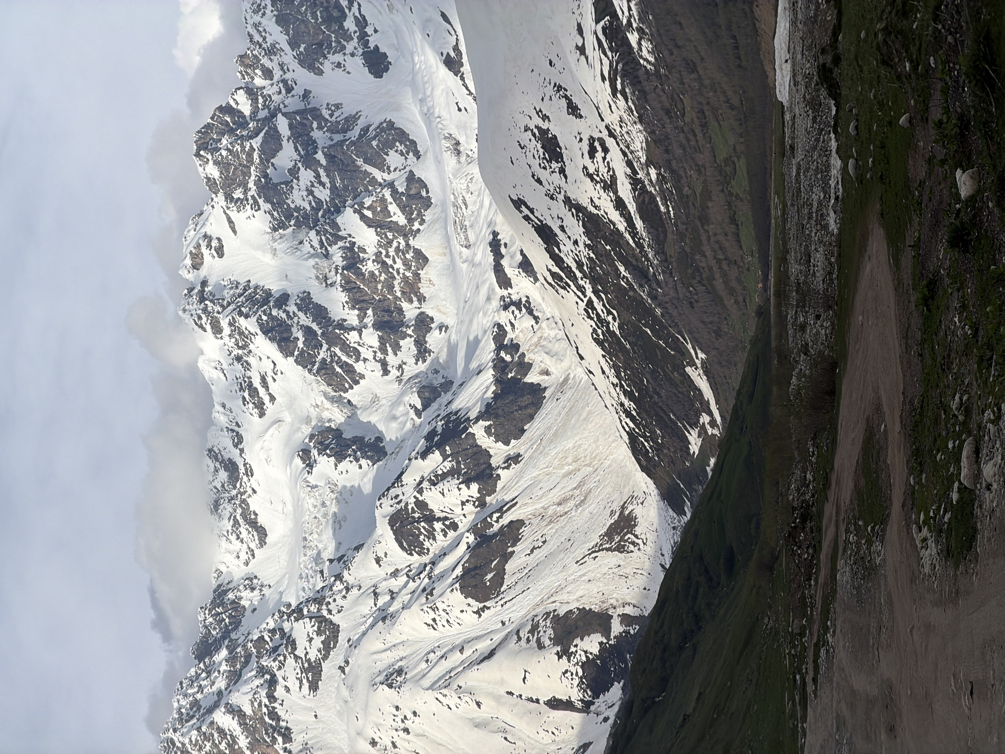 This is a vertical, medium-shot photograph of a mountain landscape in Ushguli, Georgia. In the foreground, a dirt road winds through a grassy, rocky area. The middle ground features a valley with sparse trees and a wide, rocky riverbed, partially covered by snow on the right. The background is dominated by a majestic, snow-capped mountain range with jagged peaks and glaciers. Clouds partially obscure the top of the mountains, and the sky above is overcast and light. The overall scene depicts a rugged, natural environment with evidence of a road indicating human presence in an otherwise wild setting. There are no people or animals visible in the image.