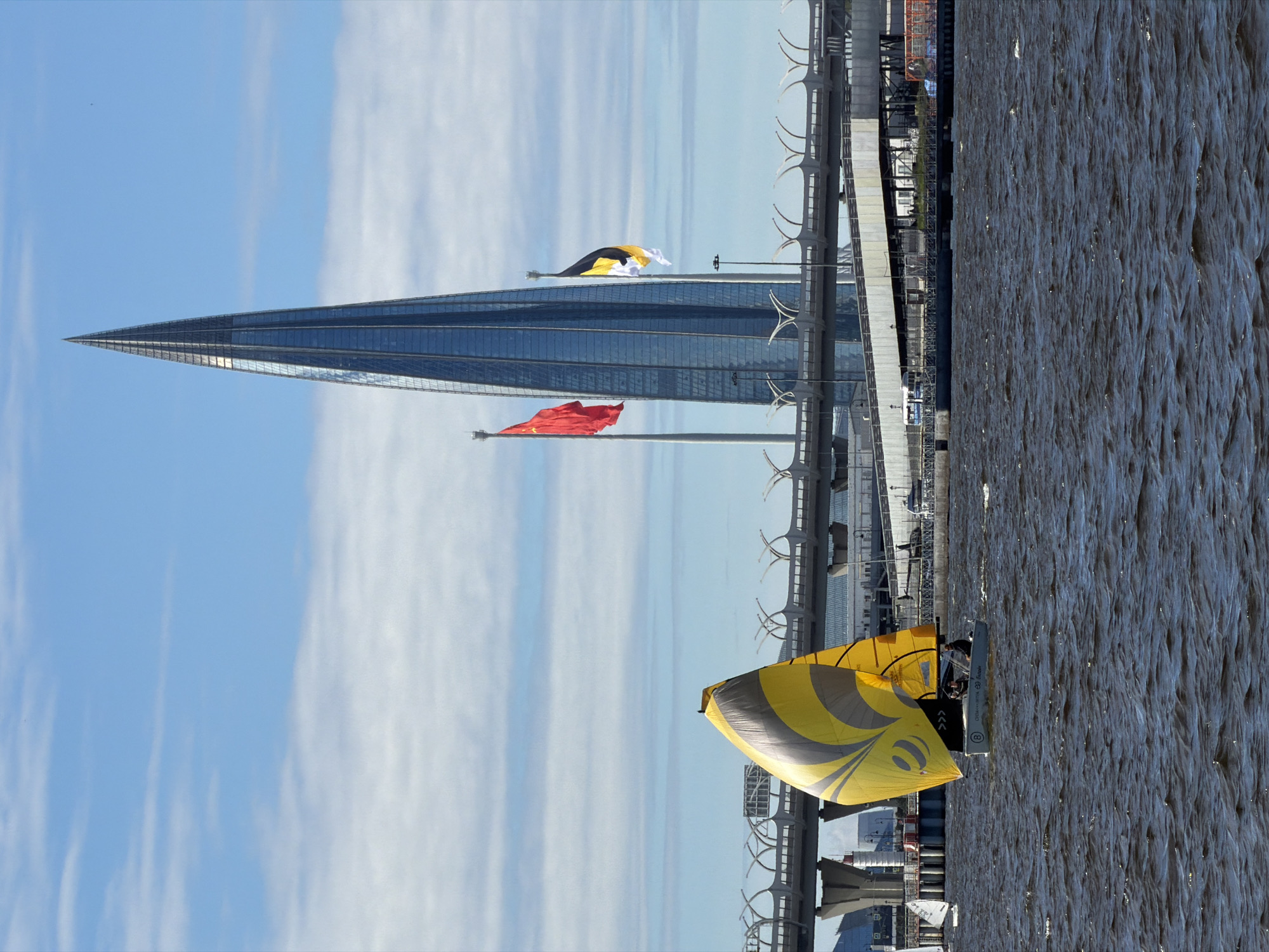 The image depicts the Lakhta Center skyscraper in Saint Petersburg, Russia, under a cloudy blue sky. In the foreground, a small sailboat with a yellow and grey sail is sailing on choppy dark water. Two people are visible on the sailboat. In the background, a multi-level highway overpass spans across the scene, with the Lakhta Center prominently visible behind it. Two flags, one red and one black and yellow, are flying from poles in front of the skyscraper. The overall scene suggests a bright, windy day on the water in an urban environment.