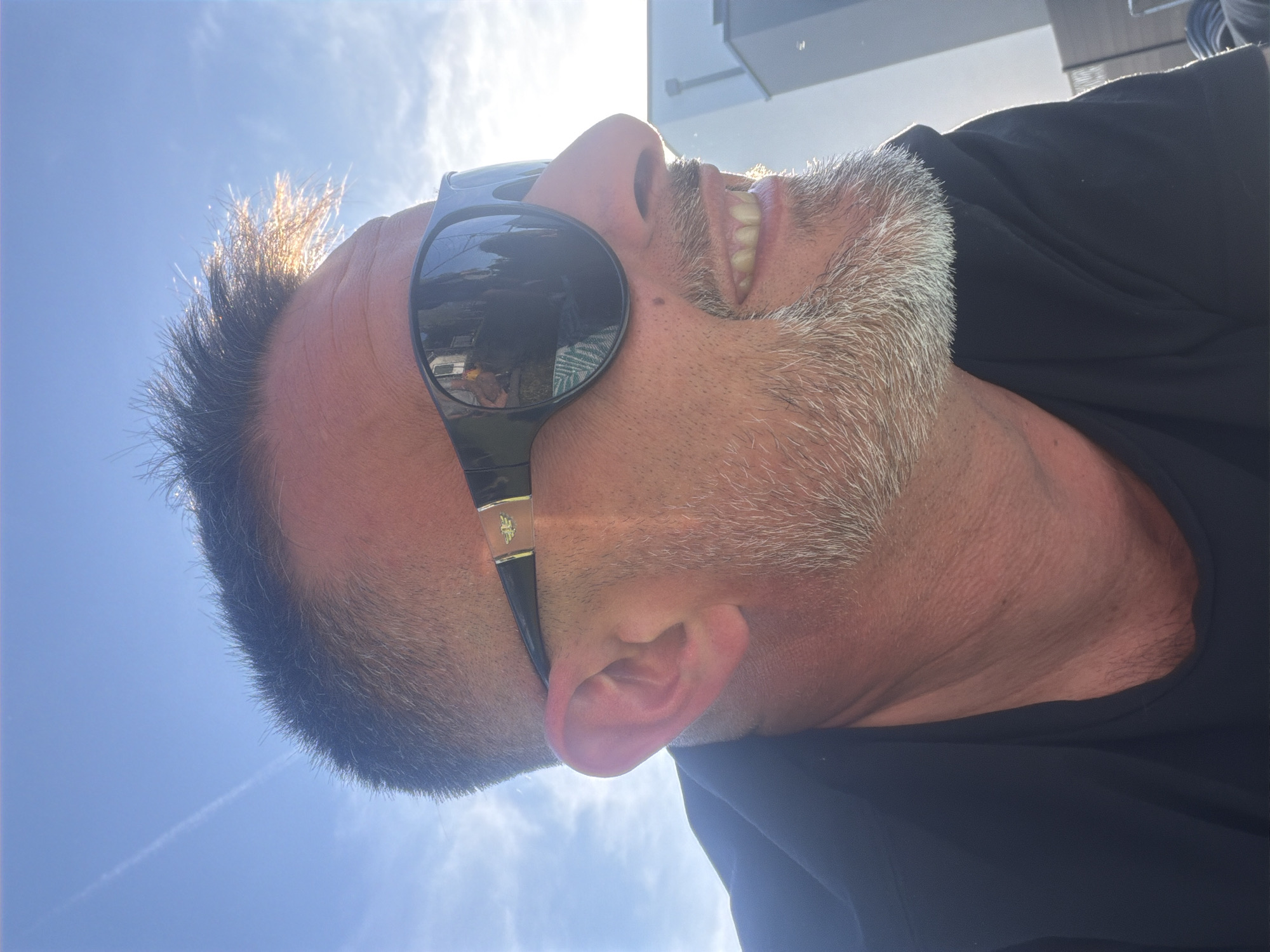 A medium shot looking up at a man wearing sunglasses. The man has short, dark hair and a gray beard. He is wearing a black shirt. The background is a bright blue sky with some white clouds. The location is most likely not Junglinster, Luxembourg, as there is no indication of the location in the image.