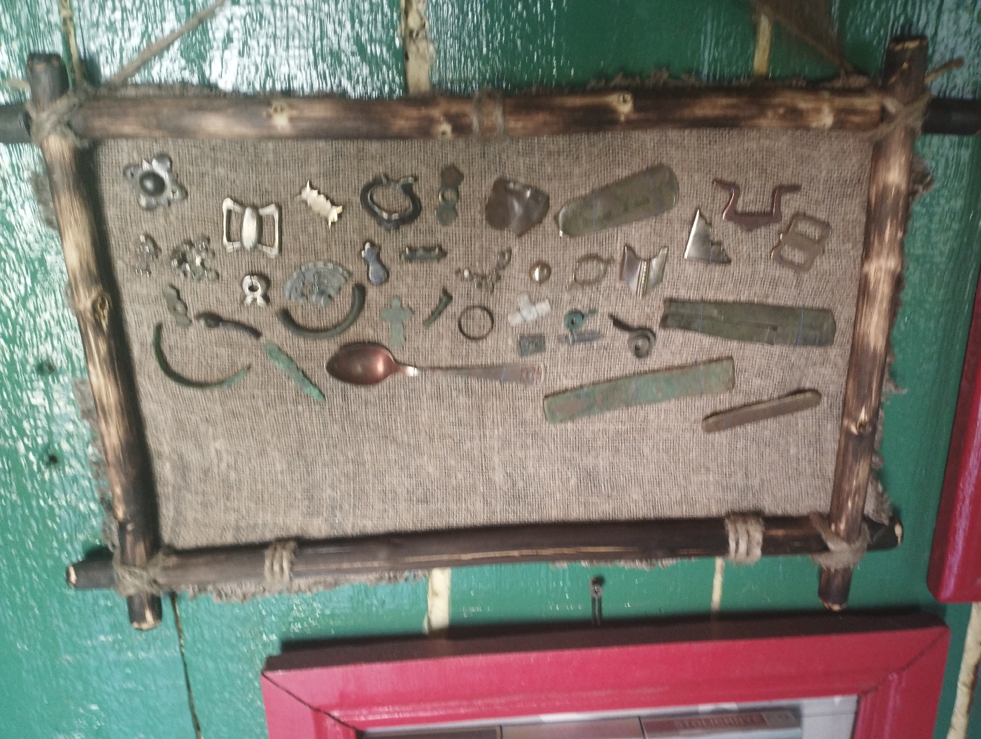 A collection of antique or historical metal objects are displayed on a beige burlap fabric stretched within a rustic wooden frame. The frame is constructed from rough, uneven branches tied together with twine. The objects, varying in size and shape, appear to be a mix of buckles, coins, small tools, a spoon, and decorative elements. Some items are heavily patinated, suggesting age and exposure.
The burlap is pinned or affixed to a dark wooden backing, which is itself attached to a green, textured surface that resembles painted wood or a painted wall. The overall presentation suggests a display or exhibit, possibly in a museum or a historical reenactment setting, showcasing artifacts. The lighting is even, and there are no clear indicators of the time of day or weather conditions. The scene is static, with no activity taking place beyond the arrangement of the objects. There is no visible text in the image. The lower portion of the image shows a red-framed structure, possibly another display or part of the room's decor, partially obscuring the view of the display.