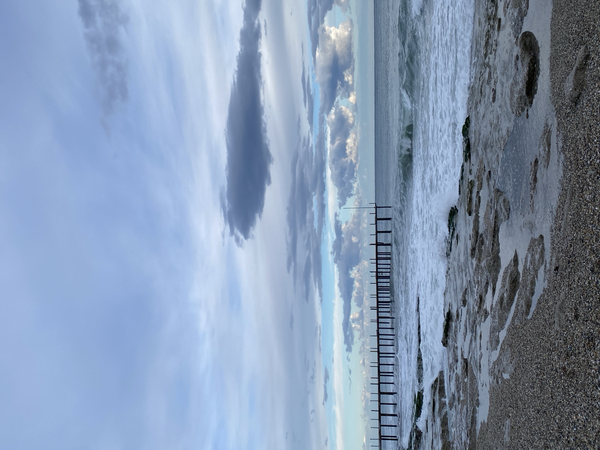 The image depicts a beach scene in Alanya, Turkey. The sky is overcast with layered clouds, ranging from light gray to dark, prominent storm clouds. The sea is moderately choppy with white foamy waves breaking along the shoreline. A series of wooden posts, possibly remnants of a pier or breakwater, stand in the water parallel to the beach. The beach consists of small pebbles and sand, with areas partially covered by water and some visible footprints. The overall lighting suggests a cloudy or overcast day.