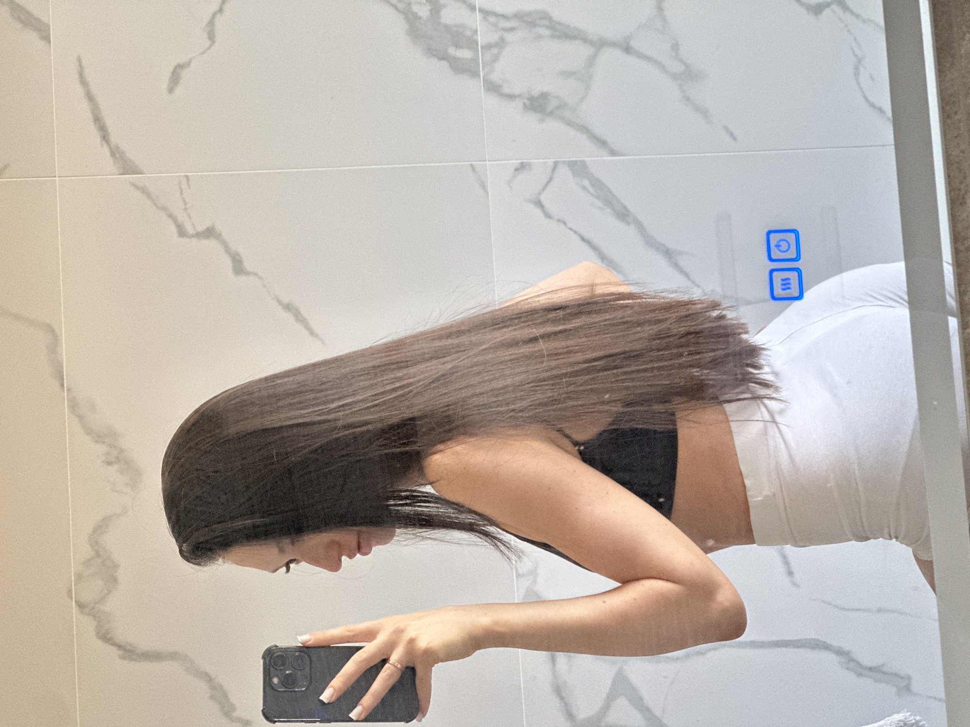 The image shows a young woman with long, dark hair taking a mirror selfie. She is wearing a black bralette and white shorts. The background appears to be a bathroom with marble-tiled walls and a lit control panel on the mirror. The woman is holding a smartphone in her left hand. There is no indication of the city of Estachi, Russia, in the image.