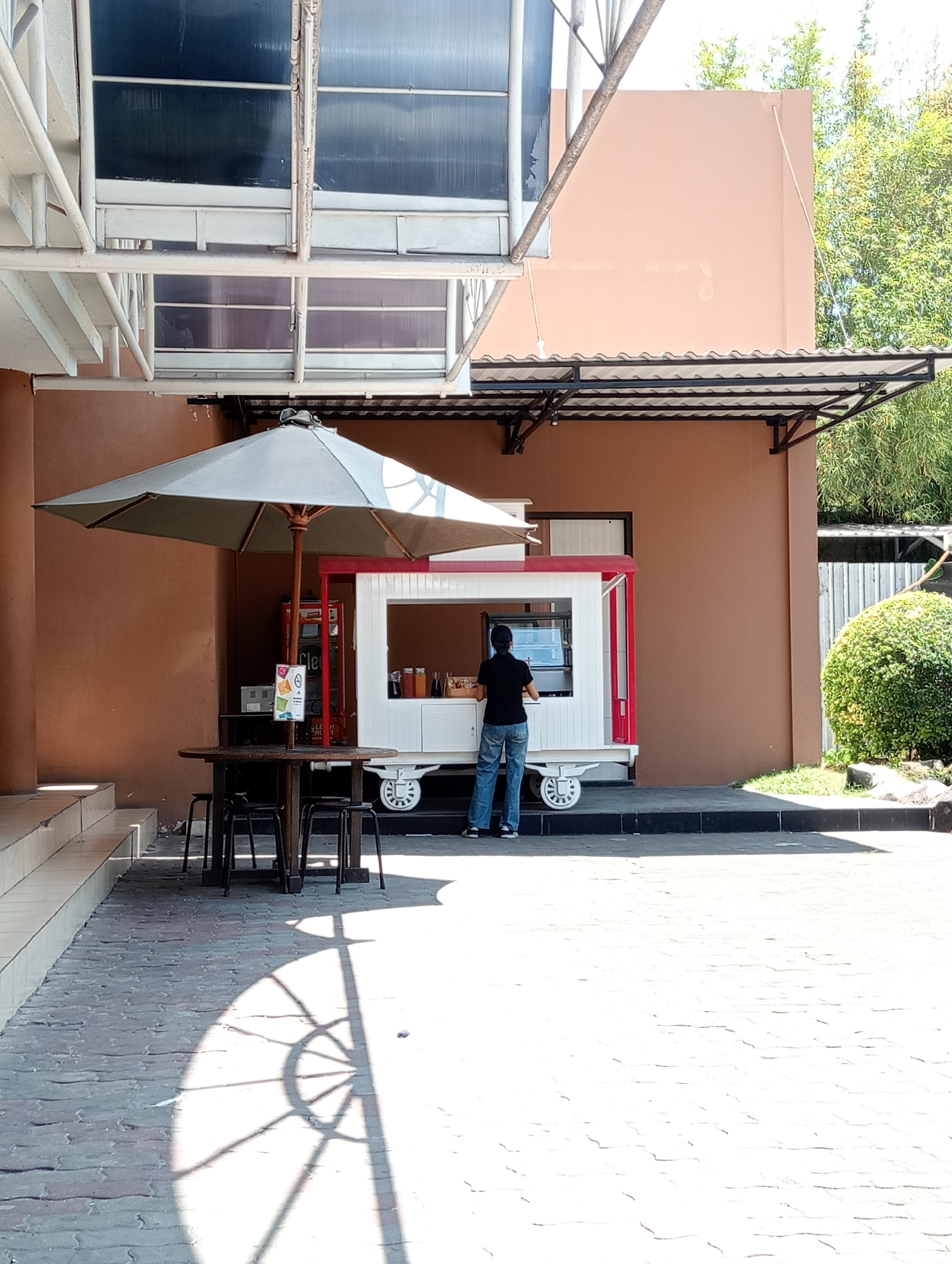 A person stands behind a white food cart with red trim in front of a brown building. The food cart has wheels and appears to be in an outdoor seating area. A large umbrella shades a round table with three black stools. The ground is paved with rectangular bricks. The building has an overhang with a metal frame and a corrugated roof. Trees are visible in the background. The scene is lit by sunlight, casting strong shadows on the ground. The person is wearing a black t-shirt and blue jeans.