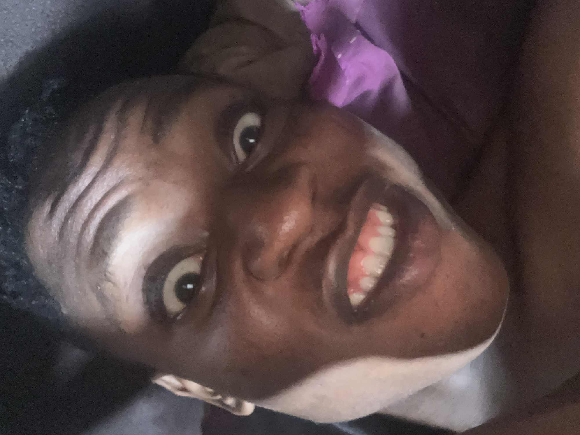 The image is a close-up, self-portrait of a person's face, taken indoors. The person has dark skin and is making a grimacing expression with their eyes wide open and mouth pulled back, revealing their teeth. The lighting is dim, casting shadows on the face and creating highlights on the skin. The background is dark and out of focus, with a hint of purple fabric visible on the right side. There is no identifiable information to suggest the location is in Ikorodu, Nigeria.