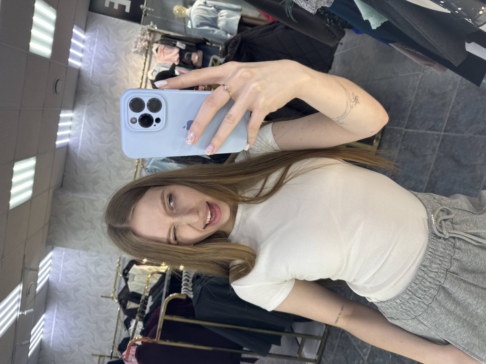A young woman is taking a selfie in what appears to be a clothing store. She is holding a blue smartphone in her right hand, showcasing a ring on her ring finger and nail art. She is winking and sticking her tongue out. She is wearing a white t-shirt and gray sweatpants. Clothing racks with various garments are visible in the background, along with a tiled ceiling with recessed lighting. Based on the metadata, the location is in the city of Chaikovsky, Russia.