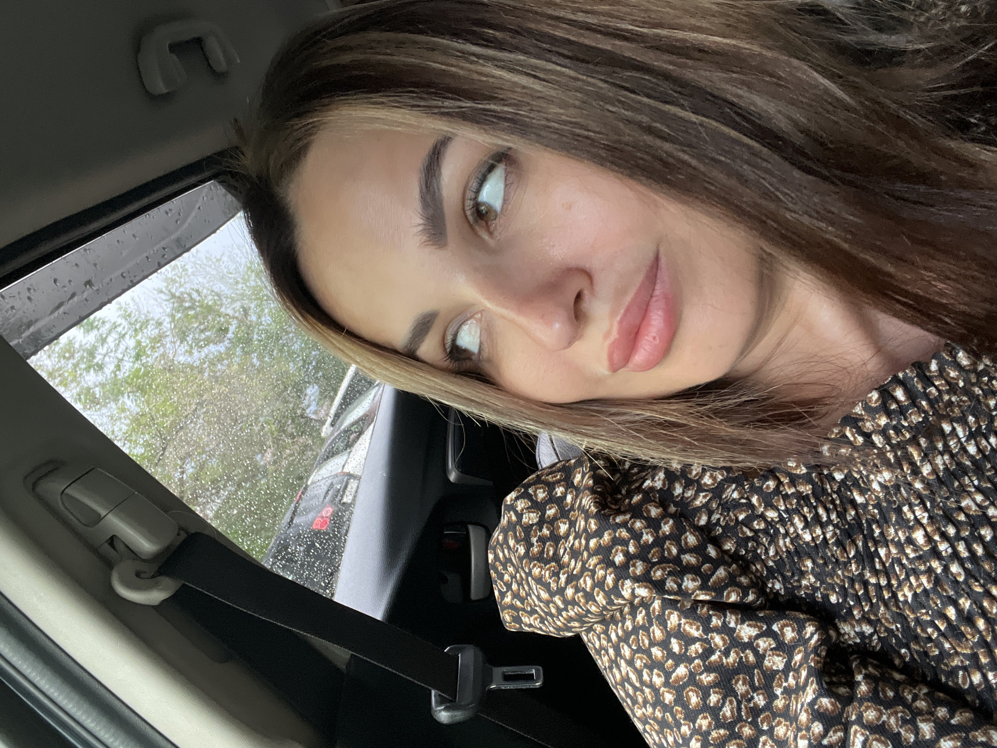 A medium shot captures a woman with brown hair streaked with lighter brown and blonde highlights, looking to her right with a slight smile. She is wearing a black dress with a brown and black leopard print pattern. Her left shoulder and arm are visible, and she appears to be in the passenger seat of a car. A black seatbelt is visible, as is the car's interior and the rainy window. The background shows other cars and trees.