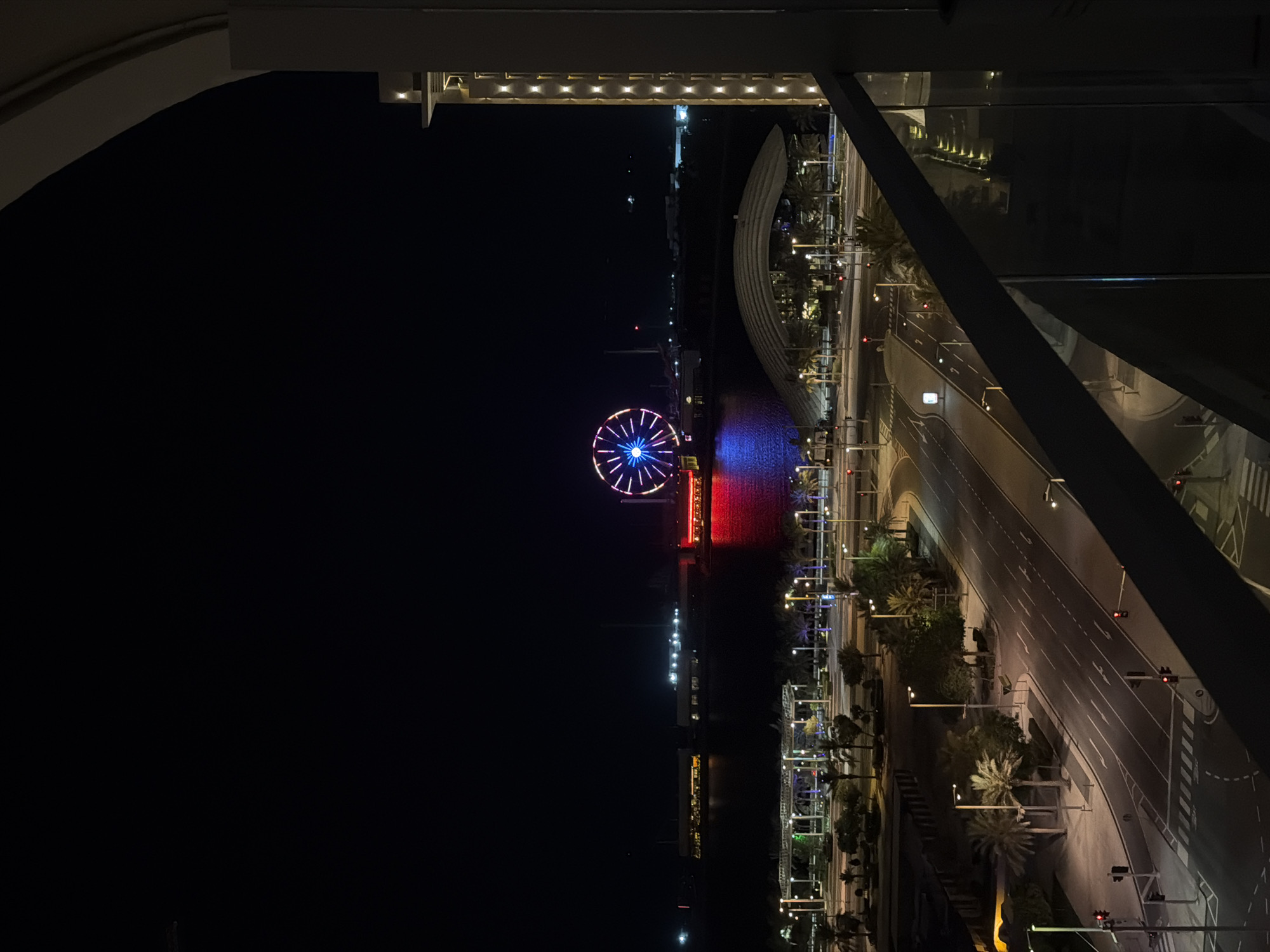 The image captures a nighttime aerial view of Lusail City, Qatar, looking towards the waterfront. Dominating the distant scene is a large, illuminated Ferris wheel, its spokes glowing with blue and white lights, reflecting on the dark water. The water itself is cast in a red glow from a nearby structure. In the middle ground, a modern building with a curved roof is visible, surrounded by palm trees and streetlights. A road with lane markings and traffic lights winds through the foreground, with palm trees lining its sides. To the right, the edge of a modern building with illuminated vertical lines is visible, suggesting a balcony or vantage point from which the photograph was taken. The sky is dark and clear, indicating it is nighttime.