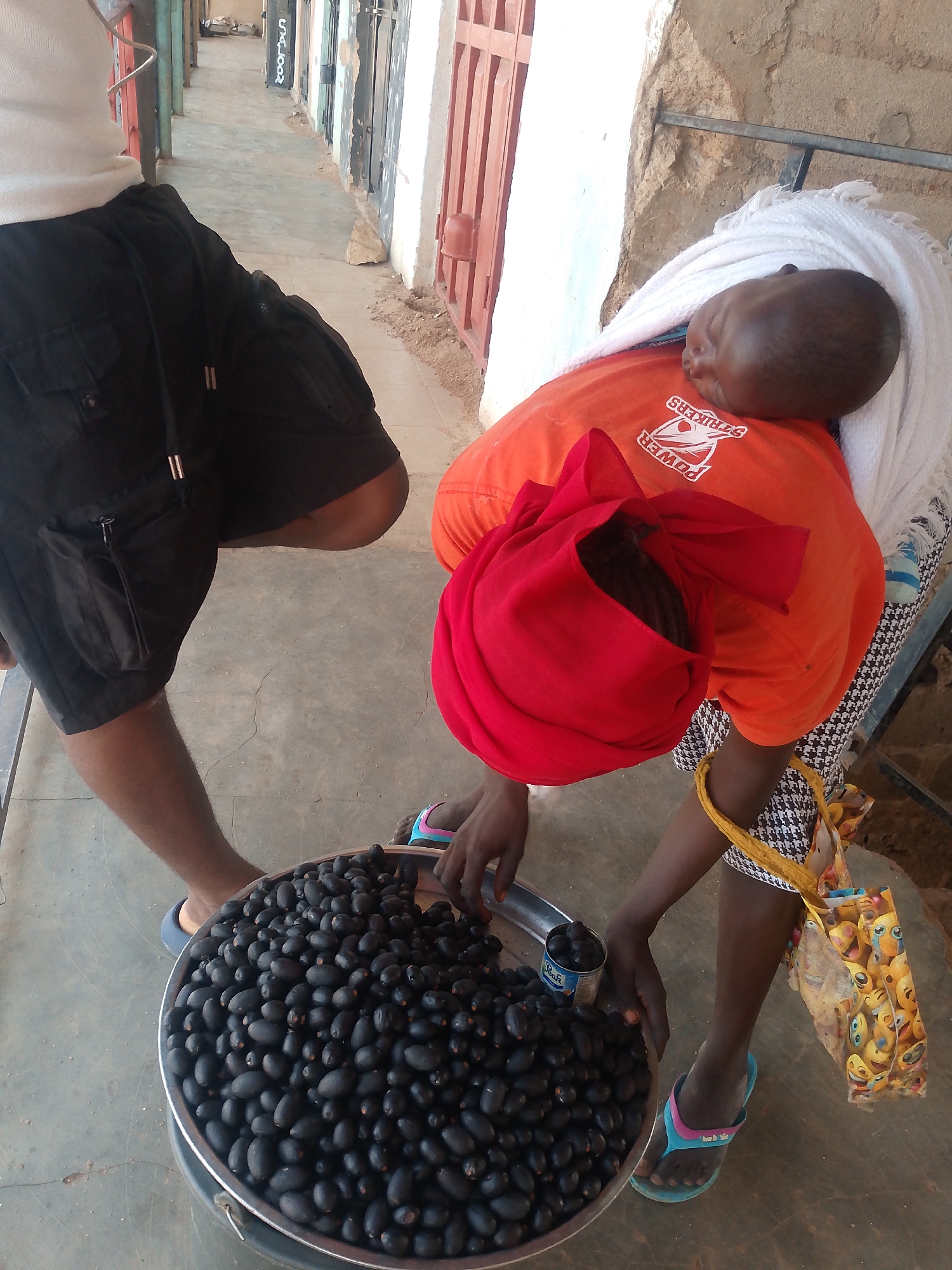 A woman in Bukuru, Nigeria, stands over a large silver basin filled with oval-shaped black fruit, possibly Java plums. She wears an orange shirt, a red headscarf, and a black and white patterned skirt. A white cloth is wrapped around her shoulder, supporting a sleeping child. She is transferring fruit into a small tin can using her hands. A man in black shorts and blue sandals stands beside her, observing. The scene takes place on a concrete walkway alongside buildings with painted doors and barred windows. An emoji-patterned shopping bag hangs from the woman’s skirt.