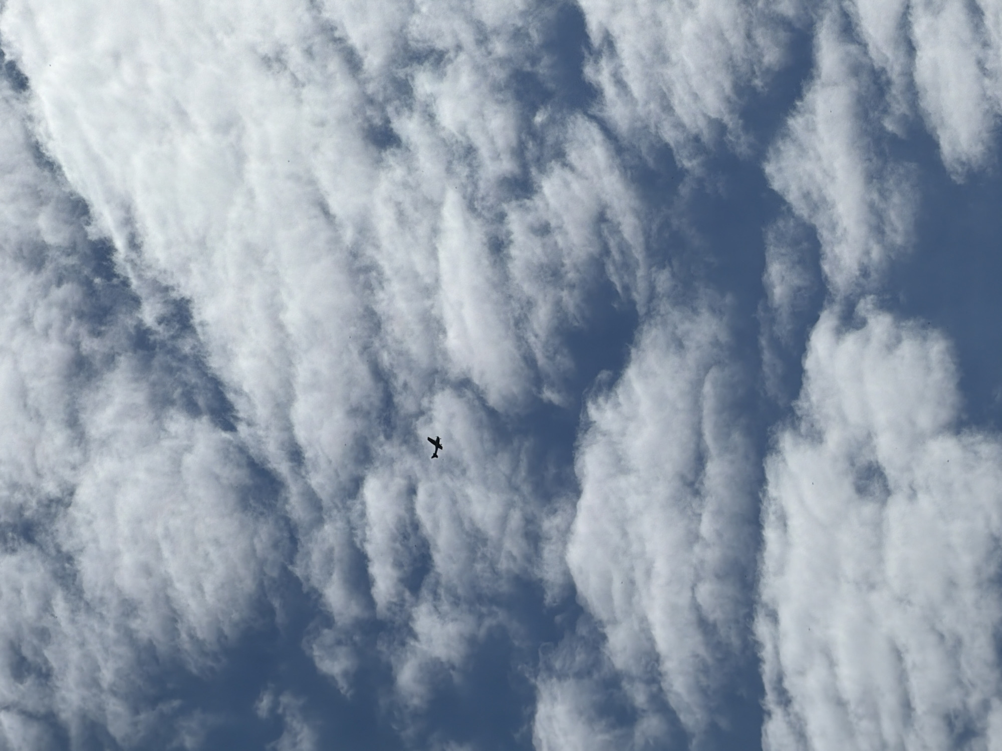 A solitary airplane is captured in silhouette against a dynamic sky filled with patterned clouds and patches of blue. The airplane, dark and small, is positioned slightly above the center of the frame, its wings and fuselage clearly visible. The clouds are predominantly white and appear to be arranged in distinct, wavy, or parallel formations, possibly altocumulus clouds. These cloud patterns create a textured background, with darker blue sky visible between the cloud bands. The lighting suggests it is daytime, with bright sunlight illuminating the clouds from above, although the sun itself is not visible. There are no other discernible objects, people, or text in the image. The scene conveys a sense of vastness and open air.