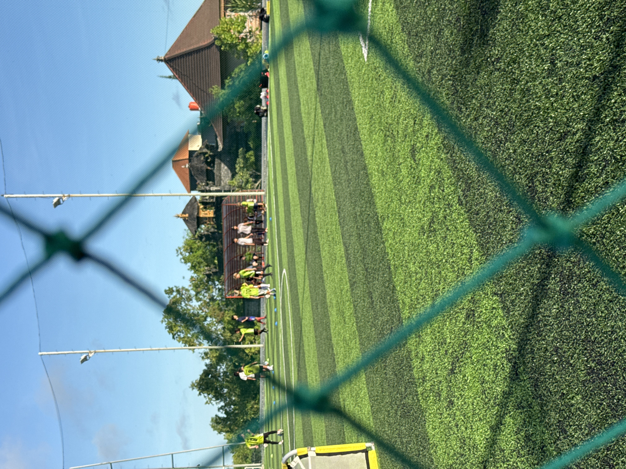 The image, taken in Kuta Selatan, Indonesia, shows a view through a green net onto an artificial turf soccer field. The field has alternating stripes of light and dark green and white boundary lines. A group of individuals wearing yellow and black athletic gear are seen in the distance, possibly practicing. A building with a brown roof is in the background, along with trees and a blue sky. White light poles are visible along the side of the field.