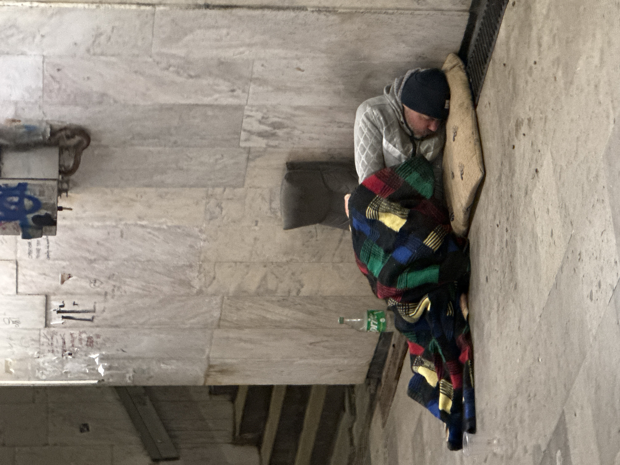 A man is sleeping on the ground, covered by a blanket. He is lying on a pad or mattress. He is wearing a grey hoodie and a dark beanie. Near him is a plastic bottle of Sprite. The location appears to be an underpass or sheltered area, with walls made of stone or marble. Graffiti is visible on the left wall. The exact location in Varna, Bulgaria is not identifiable.