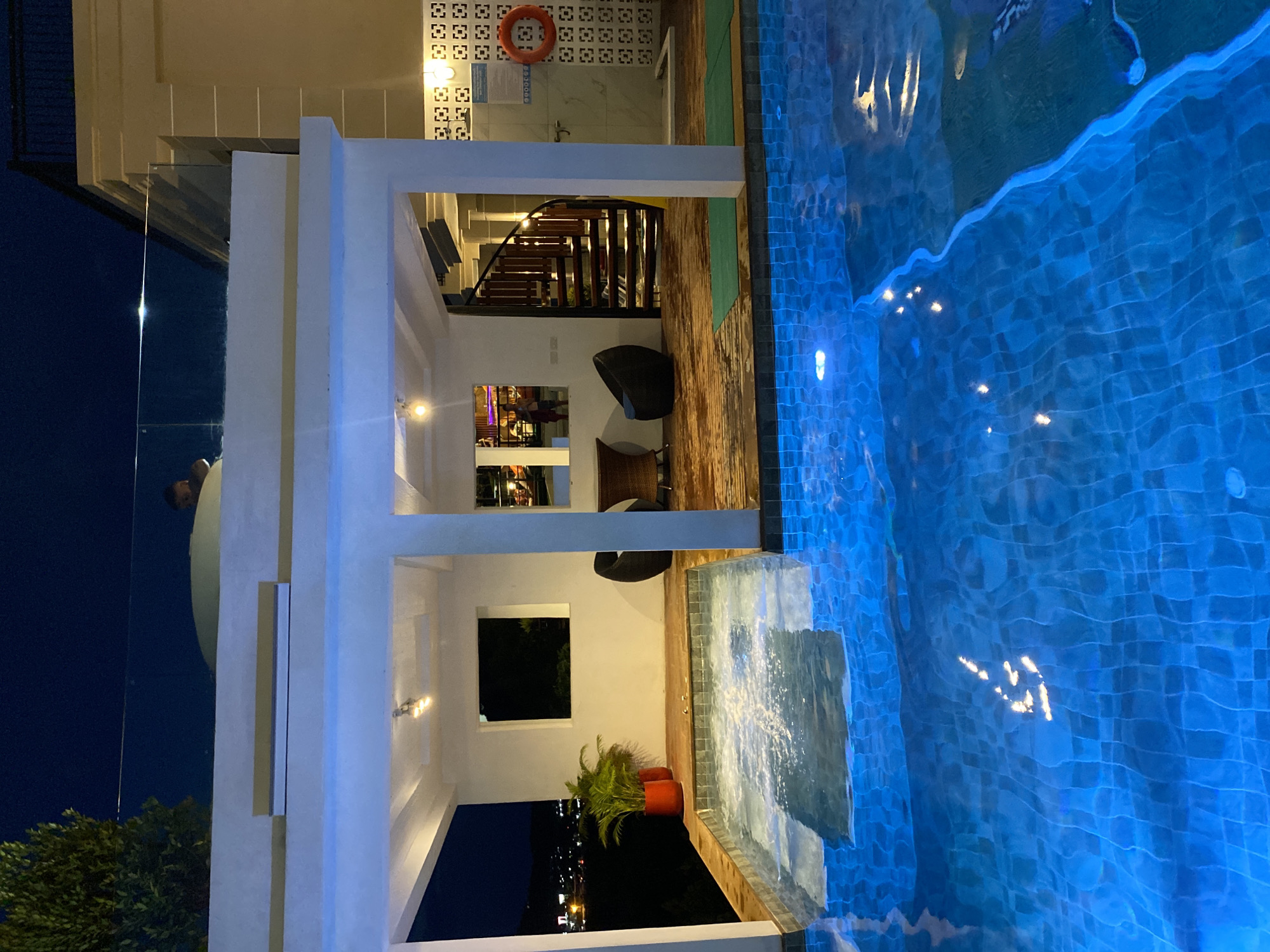 This image depicts a building with a swimming pool located in บ้านบางรักษ์, Thailand. The architecture features a white structure with a series of columns, and a glass-walled area partially obscured by a person. A wooden staircase is visible leading to an upper level. Furniture, including chairs and beanbags, are arranged within the building. There is also a potted plant near a window. The adjacent pool is filled with bright blue water and has visible tilework. In the background, the night sky can be seen, along with city lights.