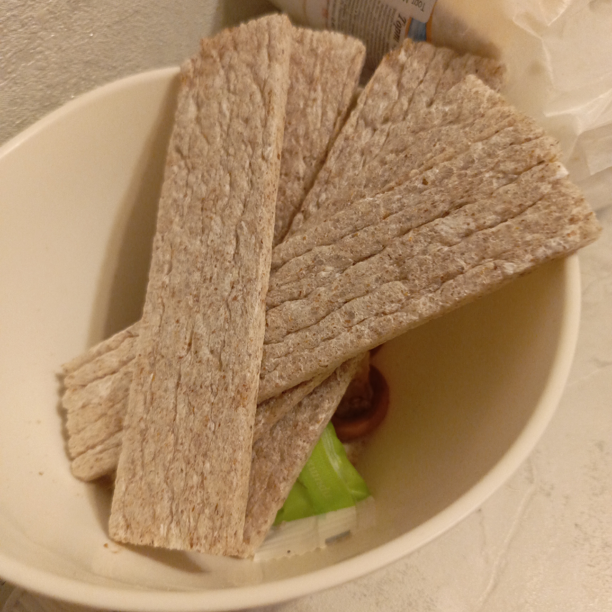A close-up shot shows a beige bowl containing several rectangular crispbreads. Some packaging, including green and white elements, is visible underneath the crispbreads. The scene is indoors and the location appears to be a home kitchen or dining area.