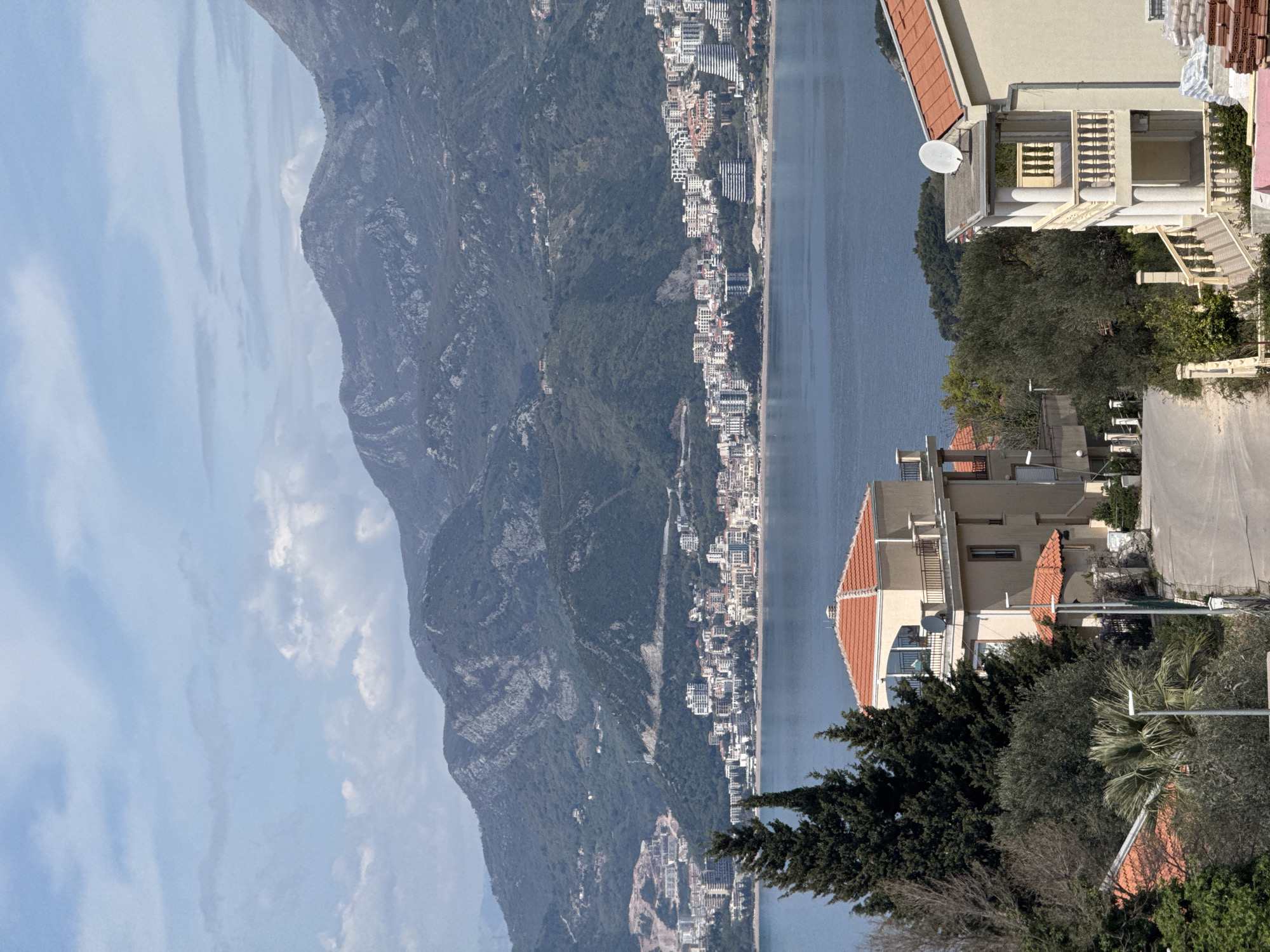 The scene depicts a coastal area in Blizikuće, Montenegro. A body of water, likely the Adriatic Sea, separates a dense mountain range from a coastal town. The town, populated with buildings of varying sizes and architectural styles, lines the coastline. On the right side of the image, foreground structures, possibly residential buildings, are visible with trees and vegetation surrounding them. A satellite dish is affixed to one of the buildings. The sky is partly cloudy.