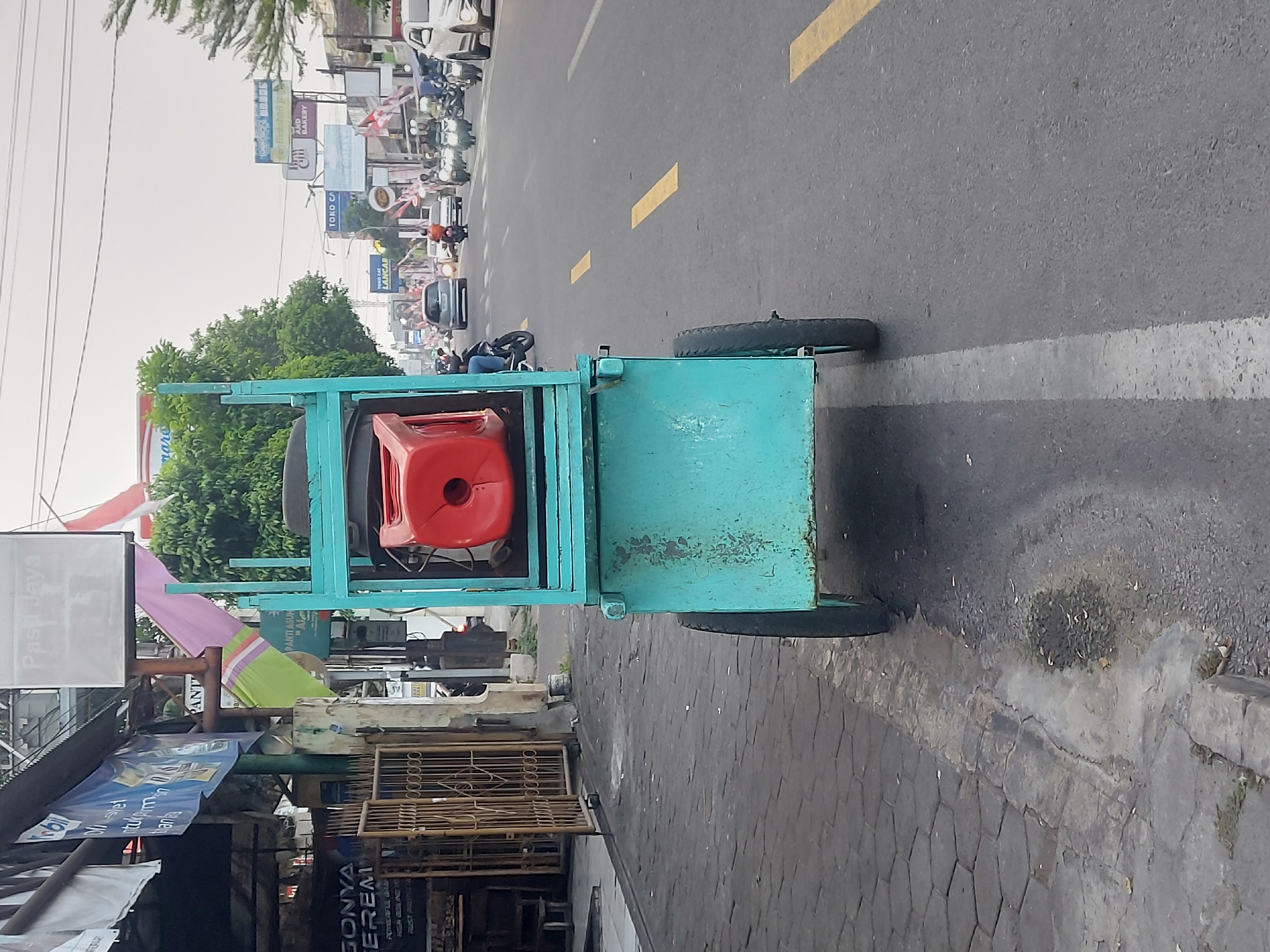 The image depicts a street scene in Yogyakarta, Indonesia, with a turquoise cart as the central subject. The cart, which appears to be a mobile vendor's setup, is positioned on the sidewalk next to a road. It has two wheels visible and holds a bright red cylindrical container on its upper platform. Behind the cart, the road is busy with traffic, including cars and numerous motorcycles. Billboards and signs are visible along the roadside, indicating commercial activity. The architecture suggests a typical urban environment in Indonesia, with shops and storefronts lining the street. The pavement on the sidewalk is made of hexagonal interlocking bricks. The overall lighting suggests it could be late afternoon or early morning.