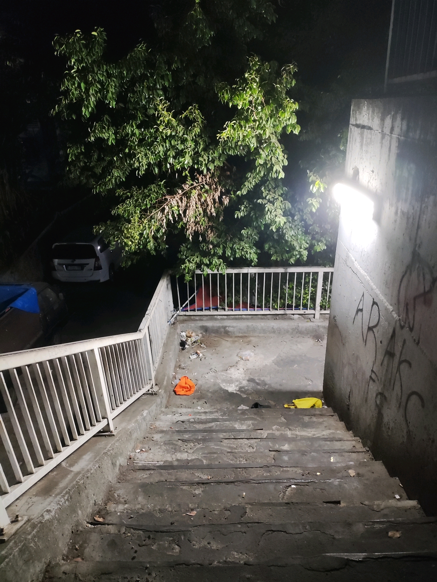 This image depicts an outdoor concrete staircase at night in East Jakarta, Indonesia. A bright floodlight illuminates the right side of the scene, casting harsh shadows. The staircase itself is worn and appears to be in disrepair, with debris scattered on its steps and landing. To the left of the staircase, a white metal railing runs along its edge and continues onto a landing. Beyond the railing, a white SUV is parked, partially obscured by lush green foliage from a tree overhead. On the landing, several items are visible, including an orange garment, a yellow bag, and other assorted trash. In the background, through the railing, a red tarp or tent-like structure is partially visible, as is a dark, indistinct area beyond. The wall to the right of the staircase is covered in graffiti.