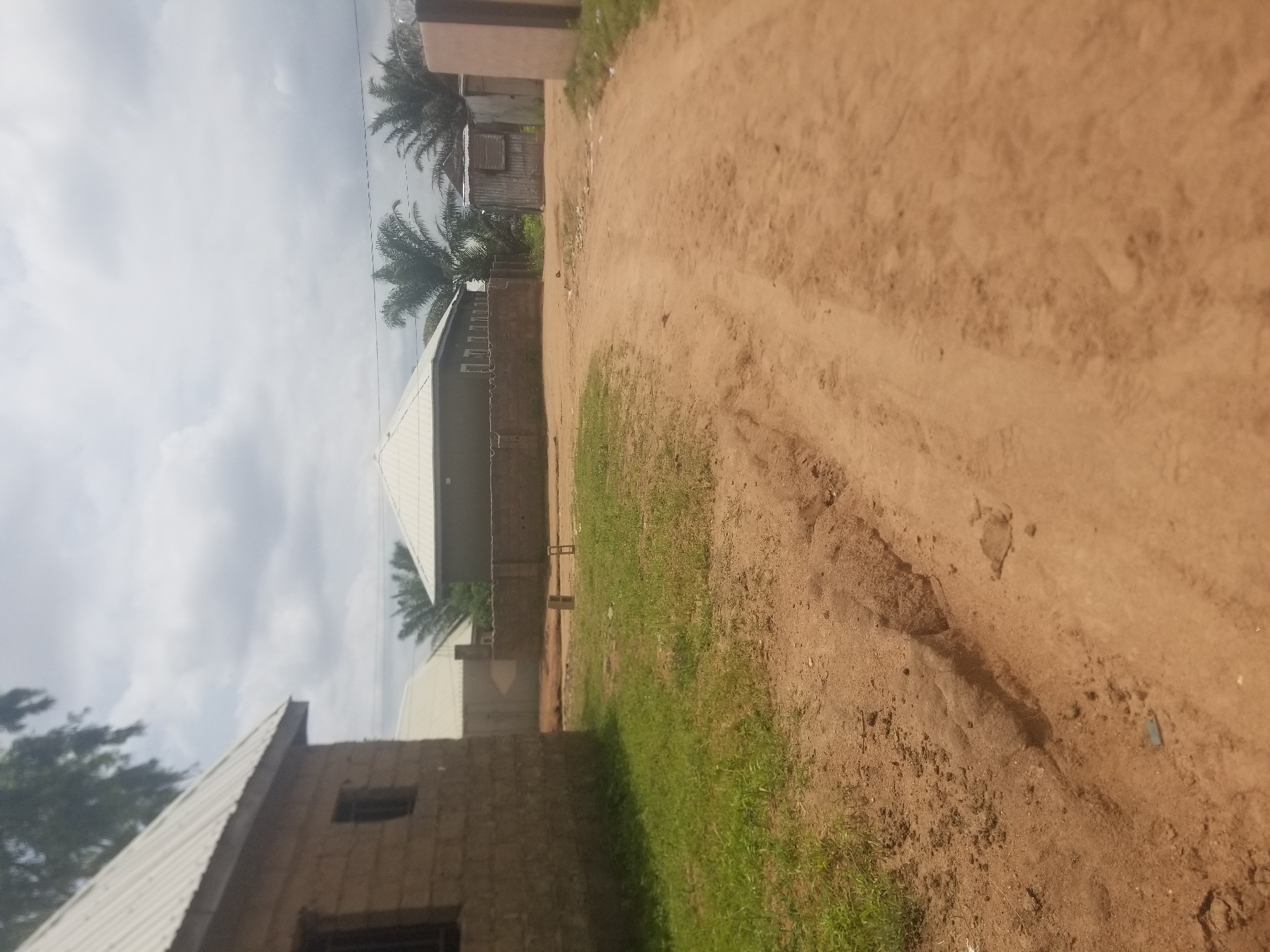 The image depicts a dirt road in Jauro Yinu, Nigeria, with a few buildings visible. On the left, a house constructed from cinder blocks with a corrugated metal roof is partially visible. Adjacent to it is another house with a similar roof. Further down the road, a walled compound encloses a building with a light-colored roof and several windows. To the right of this compound, a smaller corrugated metal structure with a window is situated. The road itself is unpaved and appears well-traveled. Vegetation, including grass and palm trees, is present around the buildings and along the road. The sky is cloudy, suggesting overcast weather. No people are present in the image.