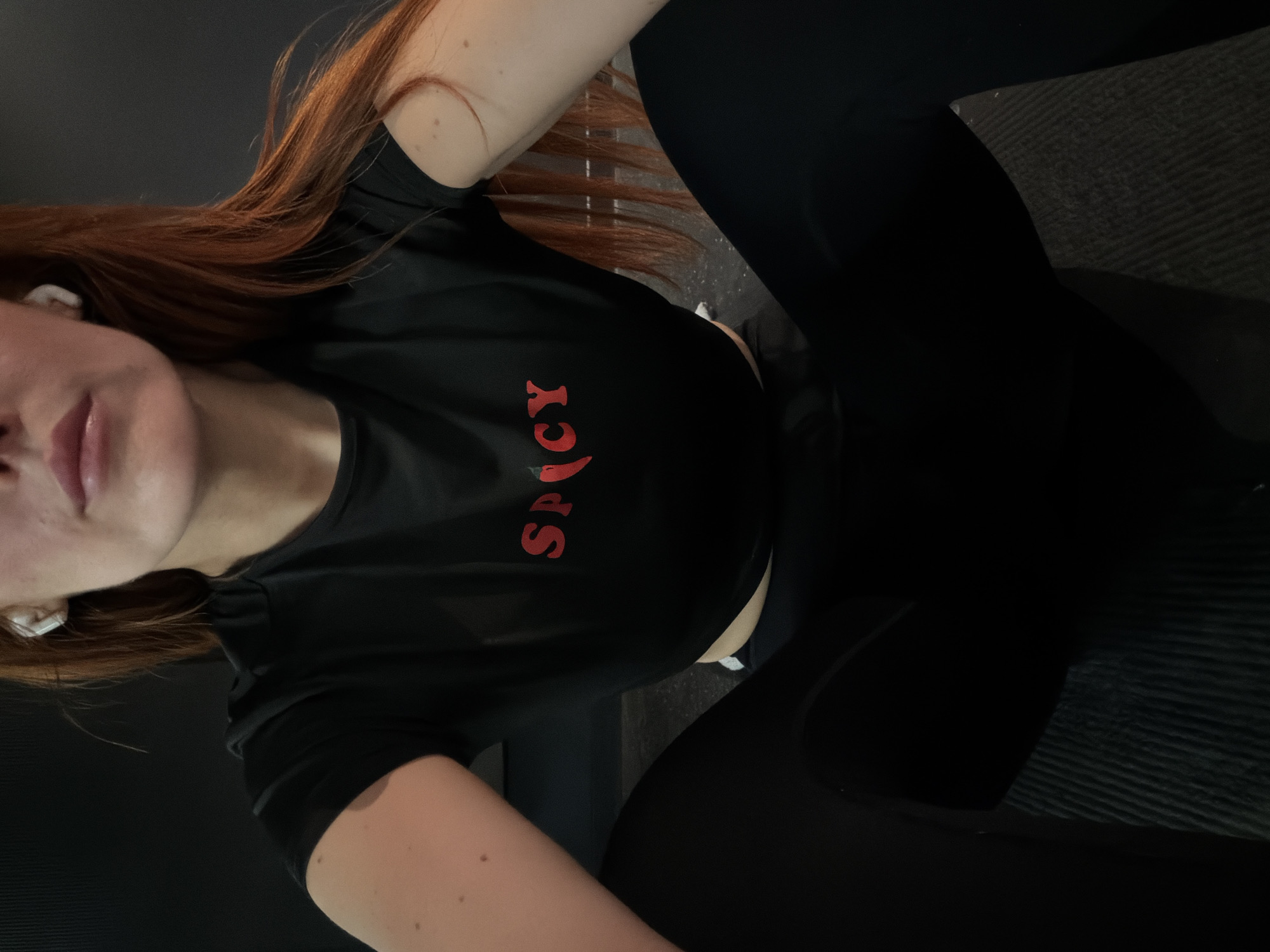 A person with red hair is wearing a black t-shirt with the word "SPICY" in red text. The person is wearing black leggings and has white earbuds in their ears. They are sitting on a dark, possibly textured surface. The location is not identifiable as Paris, France based on the visual information.