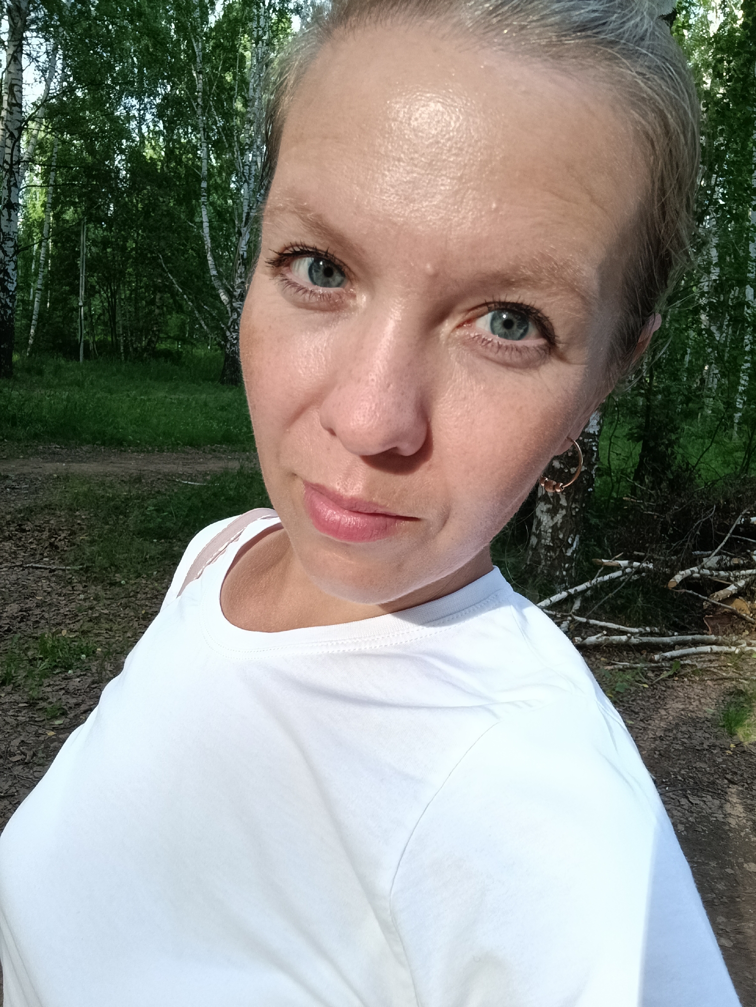 A woman with blonde hair pulled back in a bun, and blue eyes is seen in a close-up, selfie-style portrait taken outdoors. She is wearing a white t-shirt. The background consists of a dense birch forest with green foliage and scattered branches on the ground. The lighting suggests it is daytime. The image appears to be taken in a park or wooded area.