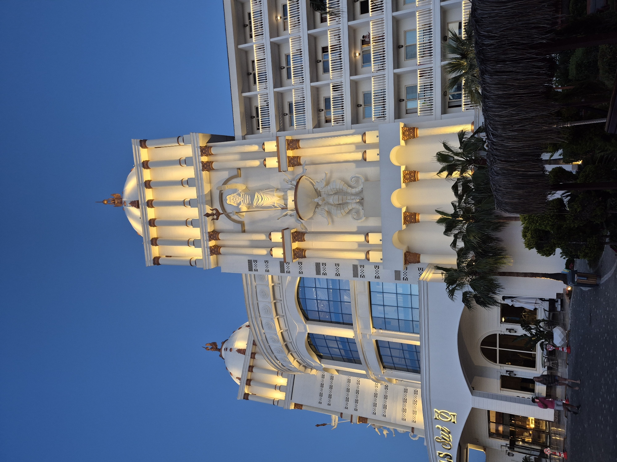 A nighttime shot shows the luxurious ASka Lara Resort & Spa hotel in Lara, Alanya, Turkey. The architecture is primarily white with gold accents, featuring Roman-style columns and sculptures. The building's facade is illuminated, contrasting against the dark blue sky. Palm trees line the front of the building, with a glimpse of the resort's entrance, where people are walking. The multi-story structure includes balconies on upper floors.