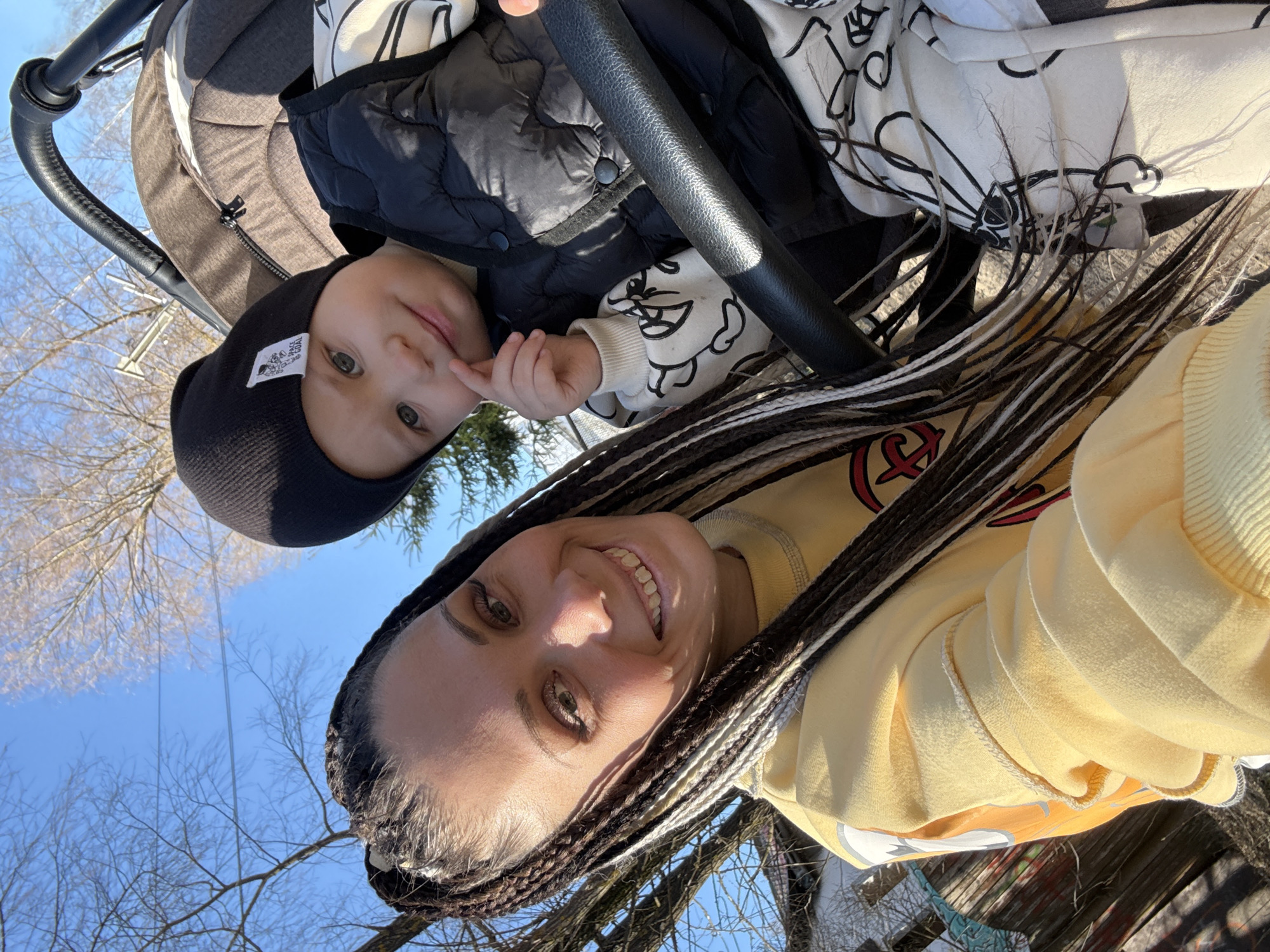 A person with braided hair, wearing a yellow sweatshirt, is taking a selfie with a baby. The baby is sitting in a stroller and is wearing a black hat, a black vest, and a white patterned long-sleeved shirt. They are both looking at the camera and smiling. The background shows a blue sky and bare trees. The location is likely in Rusanovo, Russia.