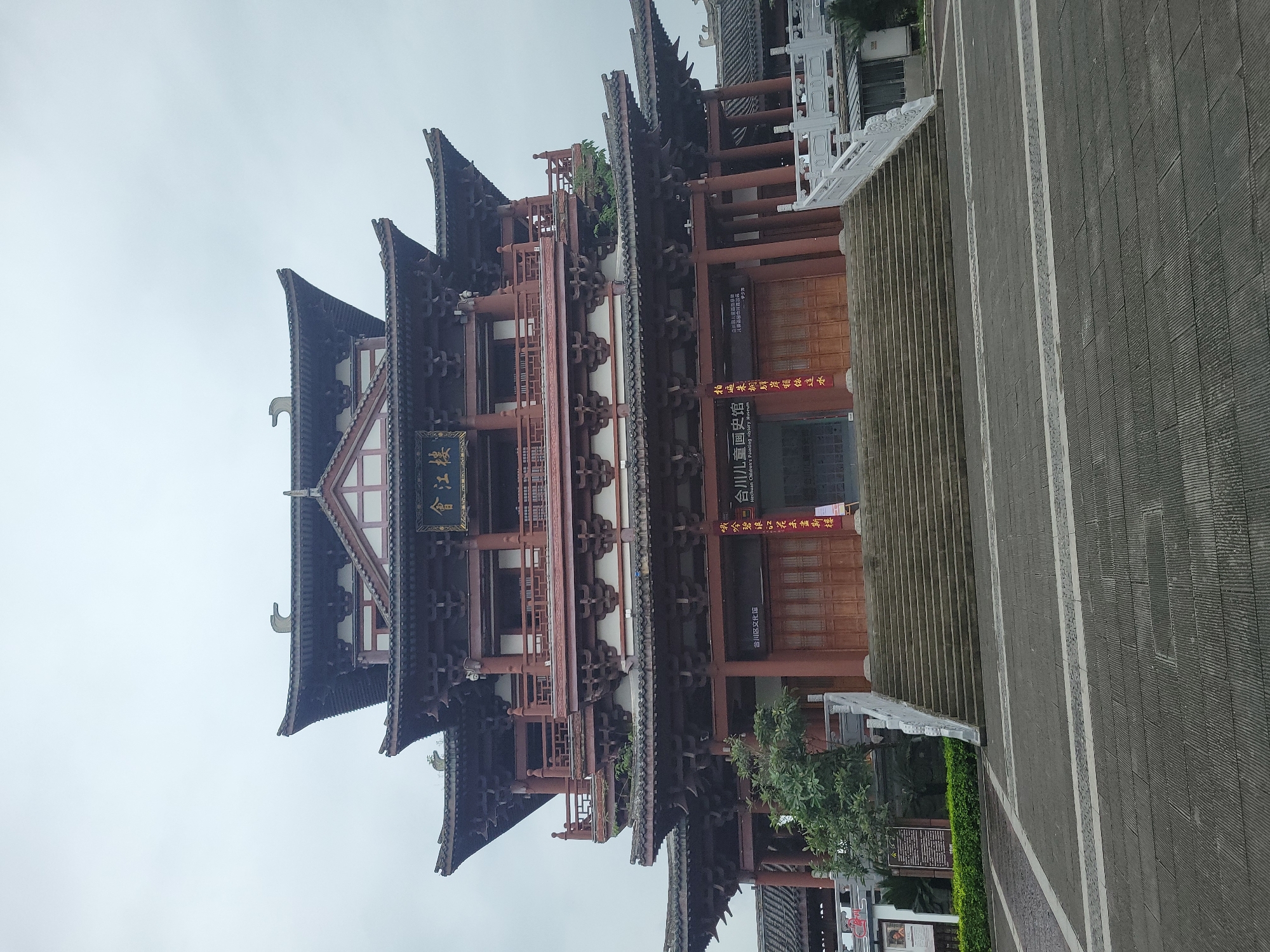 The image shows the Hejiang Tower and the Hechuan Children's Painting History Museum, which is located in the Hechuan District Cultural Center in Hechuan, China. The building features traditional Chinese architecture with multi-tiered roofs, red wooden pillars, and intricate designs. The stairs lead up to the entrance, where signs are posted in Chinese characters. The surrounding area includes paved walkways and some greenery. The sky is overcast, suggesting a cloudy day.