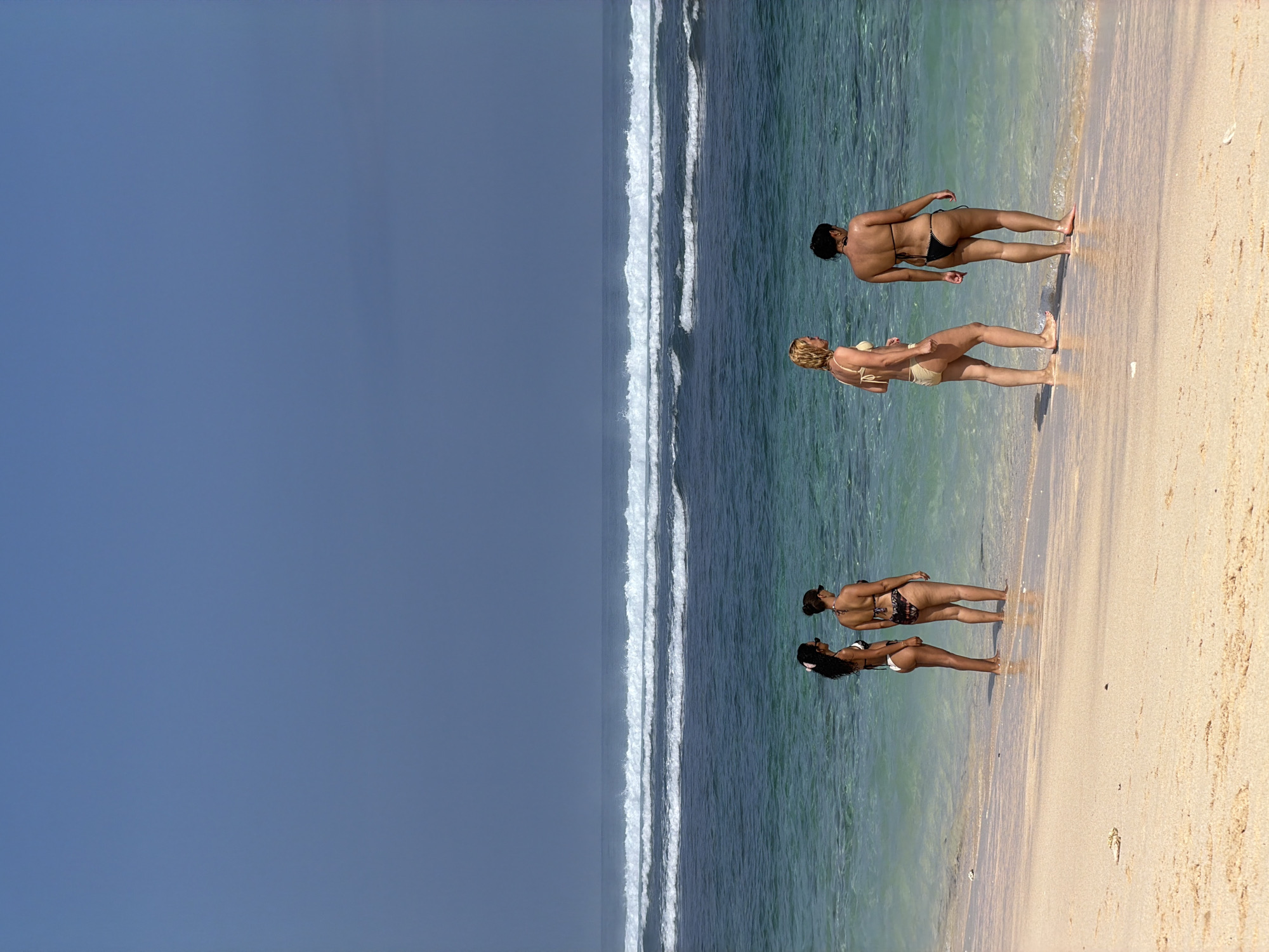 The image depicts a beach scene with four individuals in swimwear standing near the water's edge. The sky is a deep, uniform blue with a hint of gray towards the horizon. Multiple rows of white-capped waves are visible in the ocean, with the water transitioning from a light turquoise near the shore to a deeper blue further out. The sand is light-colored and appears dry.
On the left side of the image, two individuals are facing away from the camera, looking out at the ocean. The first person, closer to the water, has dark curly hair with a white clip and is wearing a white and black patterned bikini. The second person is standing slightly behind and to the right of the first, wearing a patterned bikini.
On the right side of the image, another two individuals are also facing away from the camera. The person on the left has blonde hair and is wearing a light-colored bikini. The person on the right has dark hair and is wearing a black bikini. All four individuals appear to be adults. The scene is brightly lit, suggesting daylight. The location is identified as Unknown, Unknown.