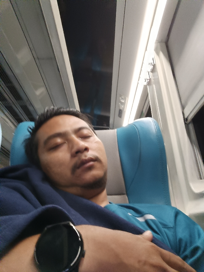An adult male is depicted resting or sleeping in a passenger seat inside a vehicle, located within the city of Purbayan, Indonesia. The subject, positioned with closed eyes and a mustache, is leaning against a light blue and gray upholstered seat. He wears a teal short-sleeved shirt, partially covered by a dark blue fabric draped over his left shoulder and chest. A black smartwatch with a round face is visible on his left wrist, with his left hand resting over his abdomen. The immediate environment includes white overhead panels with linear lighting fixtures and structural elements consistent with a train, bus, or aircraft cabin. A window frame to the right reveals darkness outside.