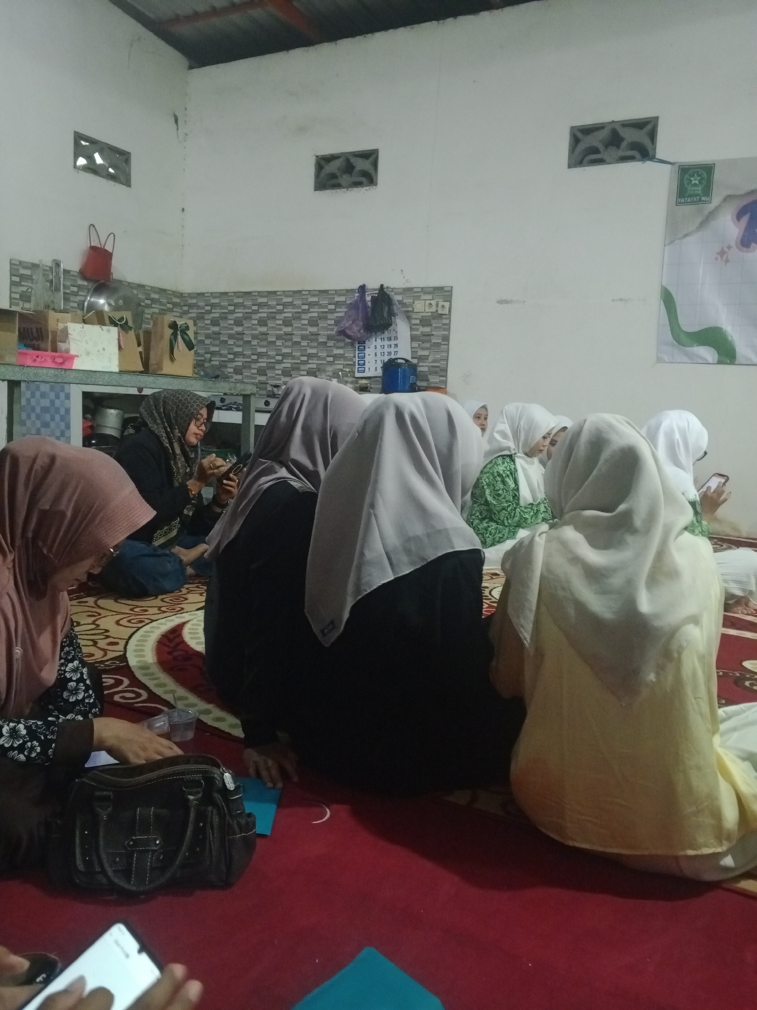 The image depicts an indoor gathering of several people, predominantly women wearing hijabs, seated on a red carpet. The environment appears to be a domestic or community space, possibly in Mlilir, Indonesia, given the presence of what appears to be a poster for "FATAYAT NU" on the wall, a prominent Indonesian Islamic organization.
In the foreground, a woman with a pink hijab is visible from the side, looking down. To her right, a black handbag rests on the carpet. Several other individuals are seated behind her, some with their backs to the camera, wearing hijabs in various colors, including gray, white, and light yellow. One woman, seated further back, is wearing a green and white patterned outfit and is looking at her phone. Another woman in a patterned hijab and glasses is seated in the middle ground, also looking at her phone.
The background reveals a wall with two ventilation grates. A shelf unit is present, holding several gift bags and a large metal bowl. A calendar is partially visible on the wall near the shelves. The overall lighting suggests it is daytime, but the interior is dimly lit. The scene suggests a casual social gathering or a religious event.