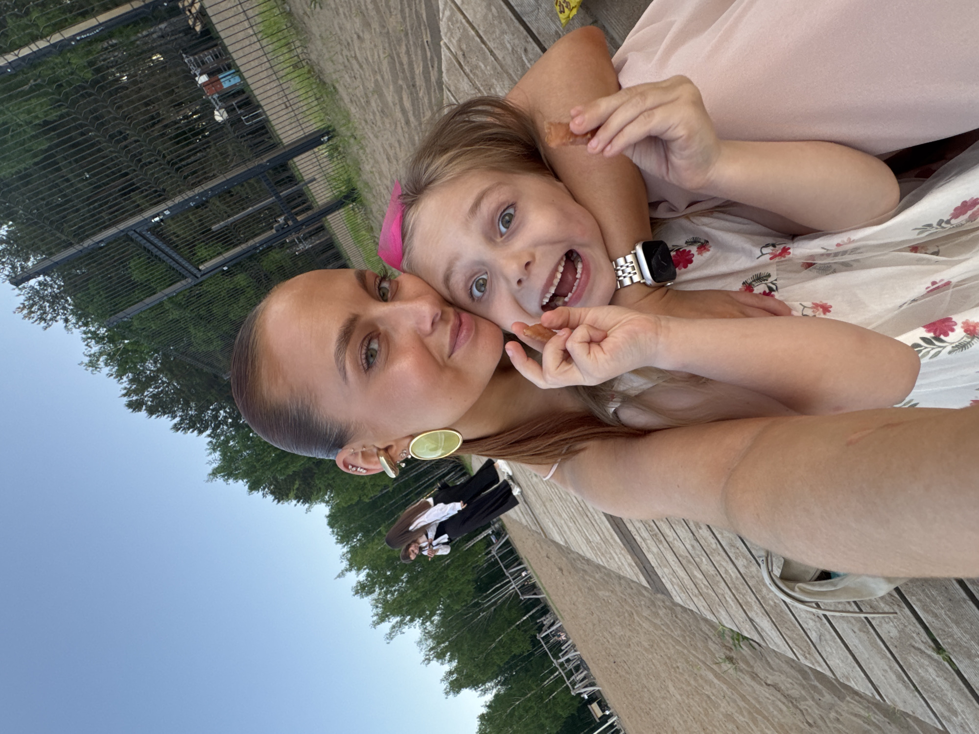 The image shows a woman and a child posing for a selfie. The woman has light brown hair pulled back, multiple ear piercings, and is wearing a large round earring. The child, with blonde hair and a pink bow, is making a silly face. Both are holding small, brown objects, possibly food. The location appears to be outdoors on a wooden deck, with a partial view of trees, a fence, and a blue sky. It's likely a park or recreational area. While the city of Chaikovsky, Russia, is mentioned, there is no forensic data to confirm this location.