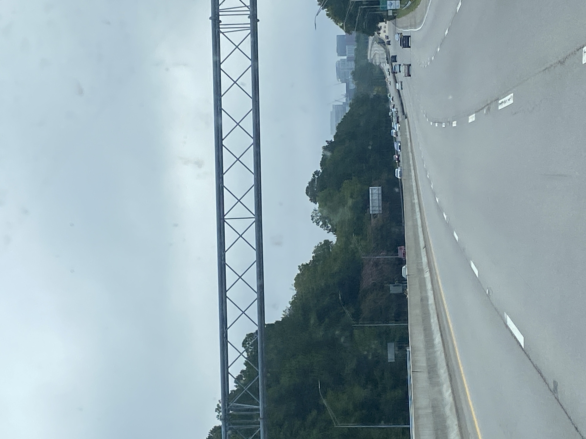 A cloudy, overcast day is depicted from the perspective of a vehicle traveling on a multi-lane highway. The highway has a concrete barrier on the left and a shoulder on the right, marked by white lines. Cars are driving in both directions on the highway. A large metal overhead sign structure spans across the highway. On the left side of the highway, a dense forest with tall green trees lines the road, and on the right side, there are more trees and some buildings visible in the distance. The city of Columbia, United States of America, is likely located in the distance where the buildings are visible.