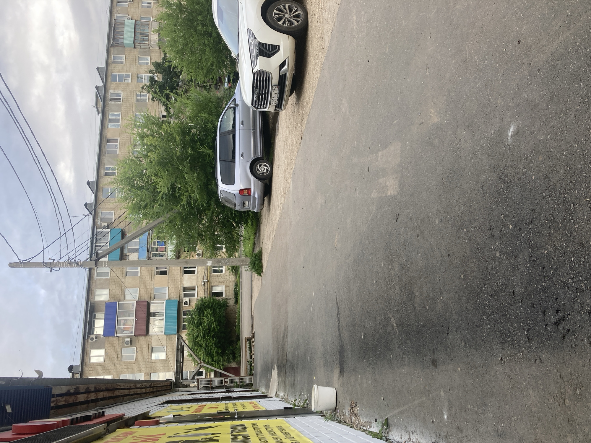 The image depicts a street scene in Balakovo, Russia, with a multi-story apartment building in the background. In the foreground, an asphalt road leads to the right, with a light-colored SUV parked on a gravel area to the right. To the left, a silver minivan is also parked on the gravel. Adjacent to the road, on the left side of the image, is the side of a building with yellow signs featuring text in Cyrillic script. A white plastic bucket is visible on the ground near this building. The sky is overcast.