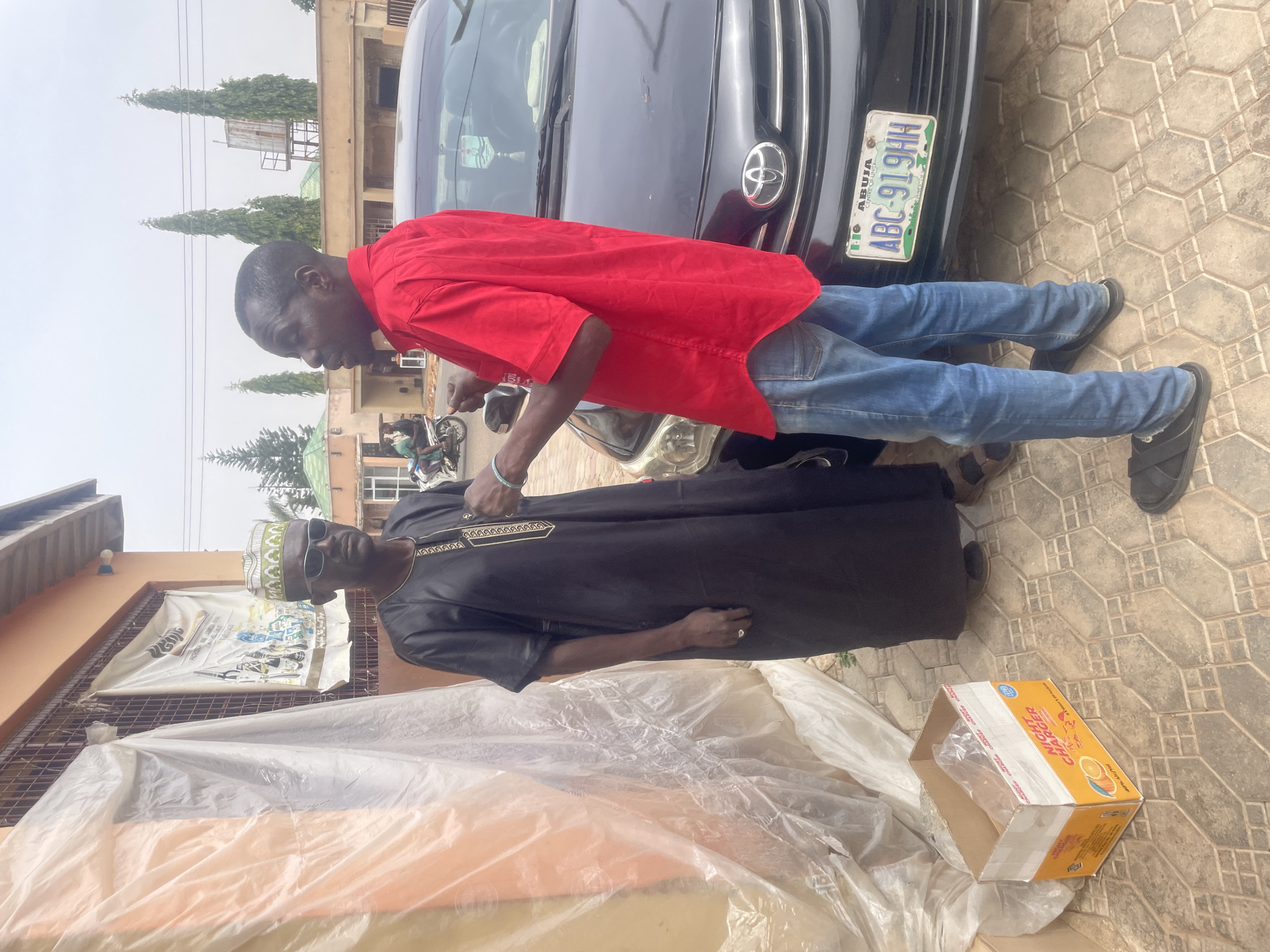 This daytime image, likely taken in Jos, Nigeria, features two men interacting outdoors on a paved surface. The sky appears overcast or hazy.
On the left, a man with dark skin wears a bright red short-sleeved shirt, blue jeans, and dark sandals. He is positioned next to a black Toyota car, which has a Nigerian license plate reading "ABUJA CENTRE OF UNITY ABC-919HH". He leans forward, seemingly engaged in conversation or a greeting with the second man.
The second man, also dark-skinned, stands to the right. He is dressed in a dark, possibly black, traditional Nigerian garment (agbada-style) with gold embroidery around the neckline, a patterned cap, and dark sunglasses.
The ground is covered with grey hexagonal interlocking paving stones. Near the feet of the man in traditional attire, there's a partially open cardboard box labeled "NIGHT CHARGER" with "40% Alc/Vol" and "PROVISUAL" visible, along with orange graphics. Clear plastic sheeting is crumpled on the ground nearby. In the background, buildings with brown facades, tall evergreen trees, and a distant water tank are visible under the hazy sky. A motorcycle can also be seen further back. Another banner or bag in the background partially displays the word "PROVISUAL".
