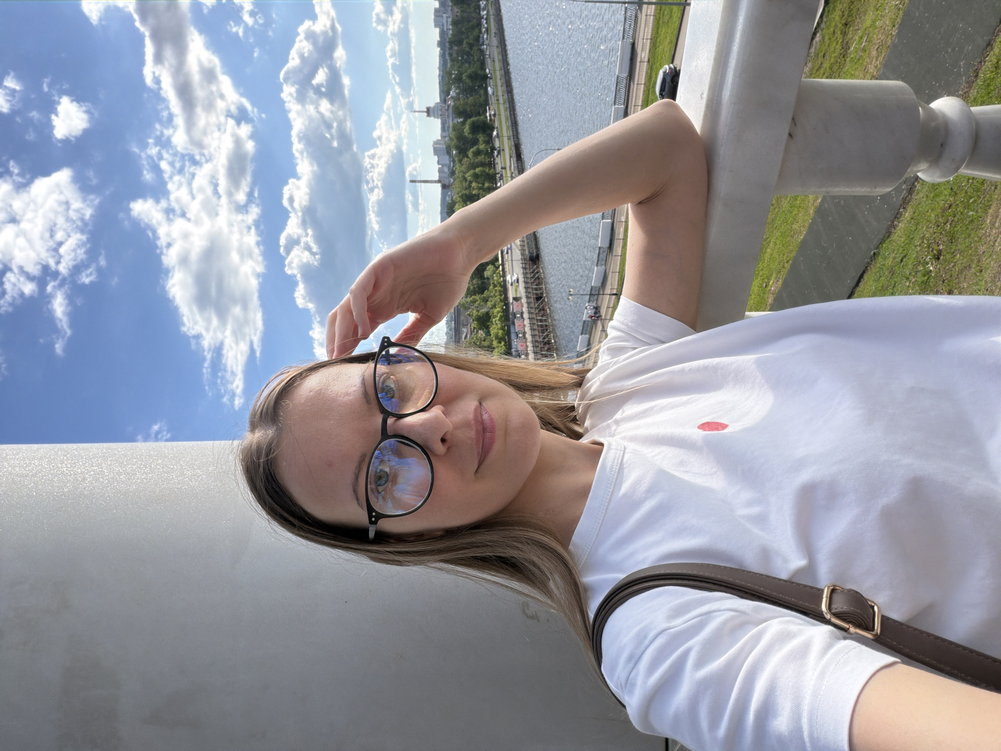A young woman is captured in a selfie-style portrait, taken outdoors on a bright, sunny day. The image is tilted, showing her head and torso, with her left arm casually raised towards her forehead and her right arm resting on a white, ornate stone railing or balustrade. She has long, light brown hair and is wearing black-framed glasses, which reflect the blue sky and white clouds. Her attire consists of a simple white t-shirt, featuring a small red, possibly heart-shaped or lip-print, detail on the left chest. A brown shoulder bag with a golden buckle is visible across her body. Her expression is neutral to slightly smiling.
The background reveals a vibrant blue sky filled with scattered white, fluffy clouds. Below the horizon, a wide body of water, likely a river or canal, stretches into the distance, bordered by a bustling urban landscape. Numerous buildings, green trees, and roads are visible along the waterfront. In the far distance, structures resembling industrial chimneys or power plant stacks can be seen. To the right of the railing, patches of green grass are visible. The scene suggests a scenic viewpoint or promenade overlooking a city and its waterway, consistent with a location like Izhevsk, Russia. There is no visible text in the image.