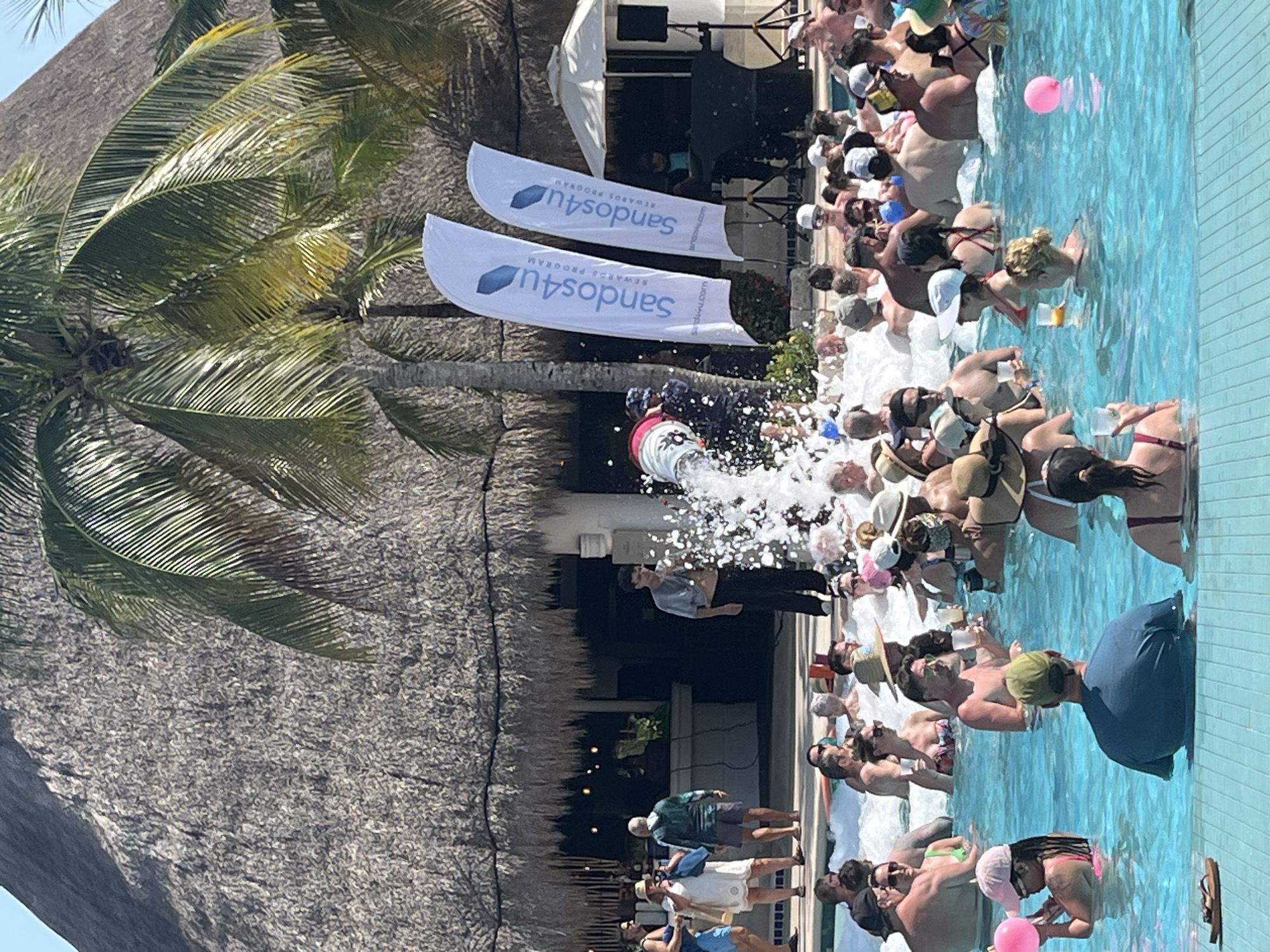 The scene is a foam party taking place at a resort in Playa del Carmen, Mexico. A crowd of people, many in swimwear, are in a pool filled with water and a thick layer of foam. A foam machine is actively spraying foam into the pool. There are two white flags with the words "sandos4u" printed on them, suggesting a sponsored event or promotional activity by Sandos Hotels & Resorts. A thatched roof structure and a palm tree are also in view, indicating a tropical resort setting. There are people standing near the pool, and some people are holding drinks in the pool.