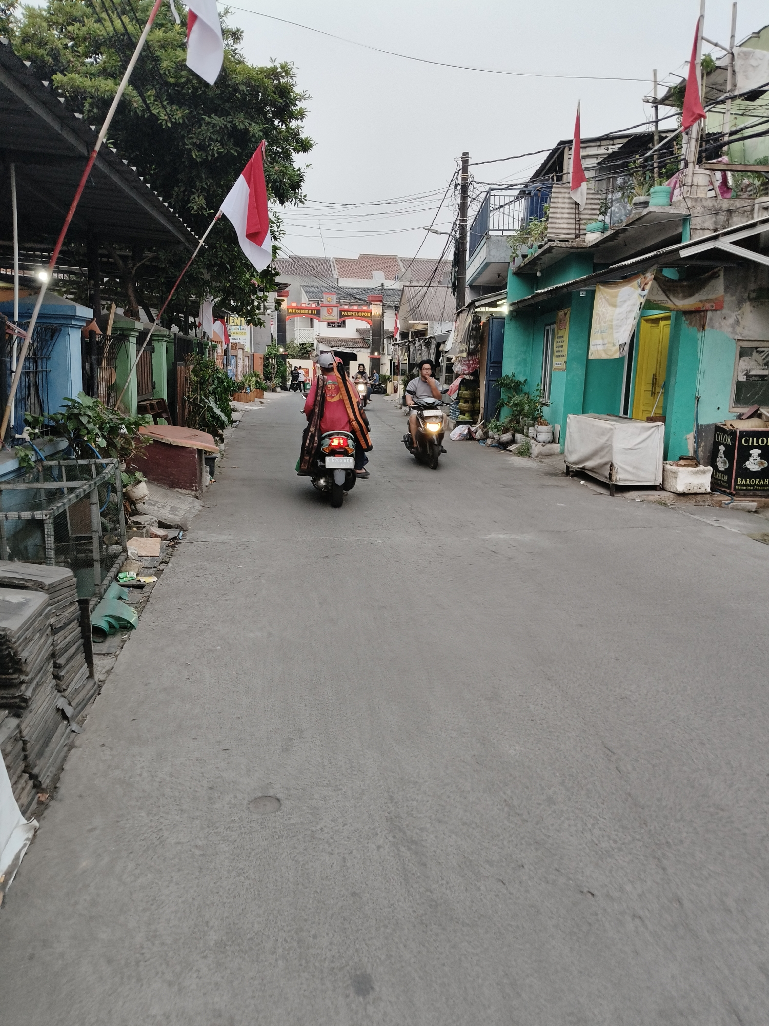 The image depicts a street scene in Ciputat, South Tangerang, Indonesia. The street is paved and appears to be an alleyway or a narrow residential road, lined with buildings of varying colors and architectural styles. Indonesian flags are prominently displayed, suggesting a national holiday or celebration.
In the foreground, on the left side of the street, there is a stack of tiles. Further down, several cages are visible, possibly for animals.
The middle of the image shows a road with two motorcycles. The first motorcycle, further away and on the right, is ridden by a person wearing a light-colored helmet and a pink shirt. They are carrying a large orange bag on their back. The second motorcycle is closer to the viewer, on the left side of the road, and is ridden by a person wearing a gray shirt and a helmet. This person is looking to the left.
In the background, a gate with signage reading "RESIMEN II PASPELOPOR" is visible, marking an entrance to a complex or area. The buildings on either side of the street are adorned with flags and decorative elements. Some buildings have balconies and intricate electrical wiring visible.
The sky is overcast, indicating a cloudy day. The overall impression is a busy, lived-in urban environment with signs of community life and national pride.