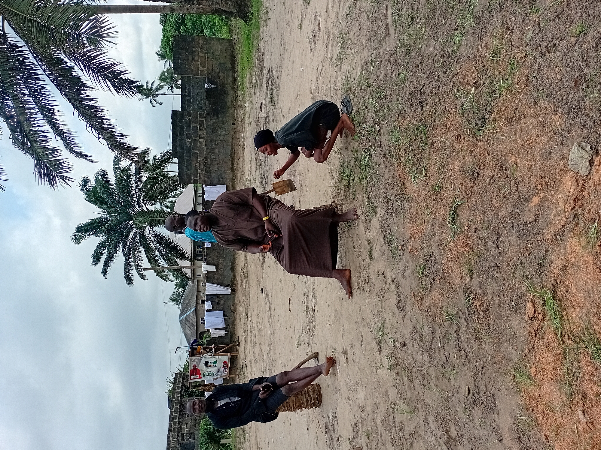 In the city of Ipota, Nigeria, four individuals are present in an outdoor, sandy area. The scene is characterized by a rough, dirt ground with sparse patches of grass and some scattered debris. A low, dark brick wall forms a partial backdrop, with laundry hanging on a clothesline in front of it. Beyond the wall, residential buildings and palm trees are visible under an overcast sky.
On the left, a young person is seated on a cylindrical object, possibly a log or a cut tree trunk. This individual wears a black jacket, shorts, and white socks, with their bare feet on the ground.
To the right of the first individual, an adult male is seated on the ground, wearing a loose, brown robe-like garment. He appears to be in conversation.
In the center, another adult male stands, partially obscured by the seated individuals. He is wearing a blue garment.
In the foreground on the right, a young person is crouched down, wearing a dark t-shirt and shorts, and a black head covering. They are holding a small, light-colored bag and appear to be actively engaged in conversation or interacting with the seated man. Their bare feet are visible. A pair of sandals lies on the ground near them.