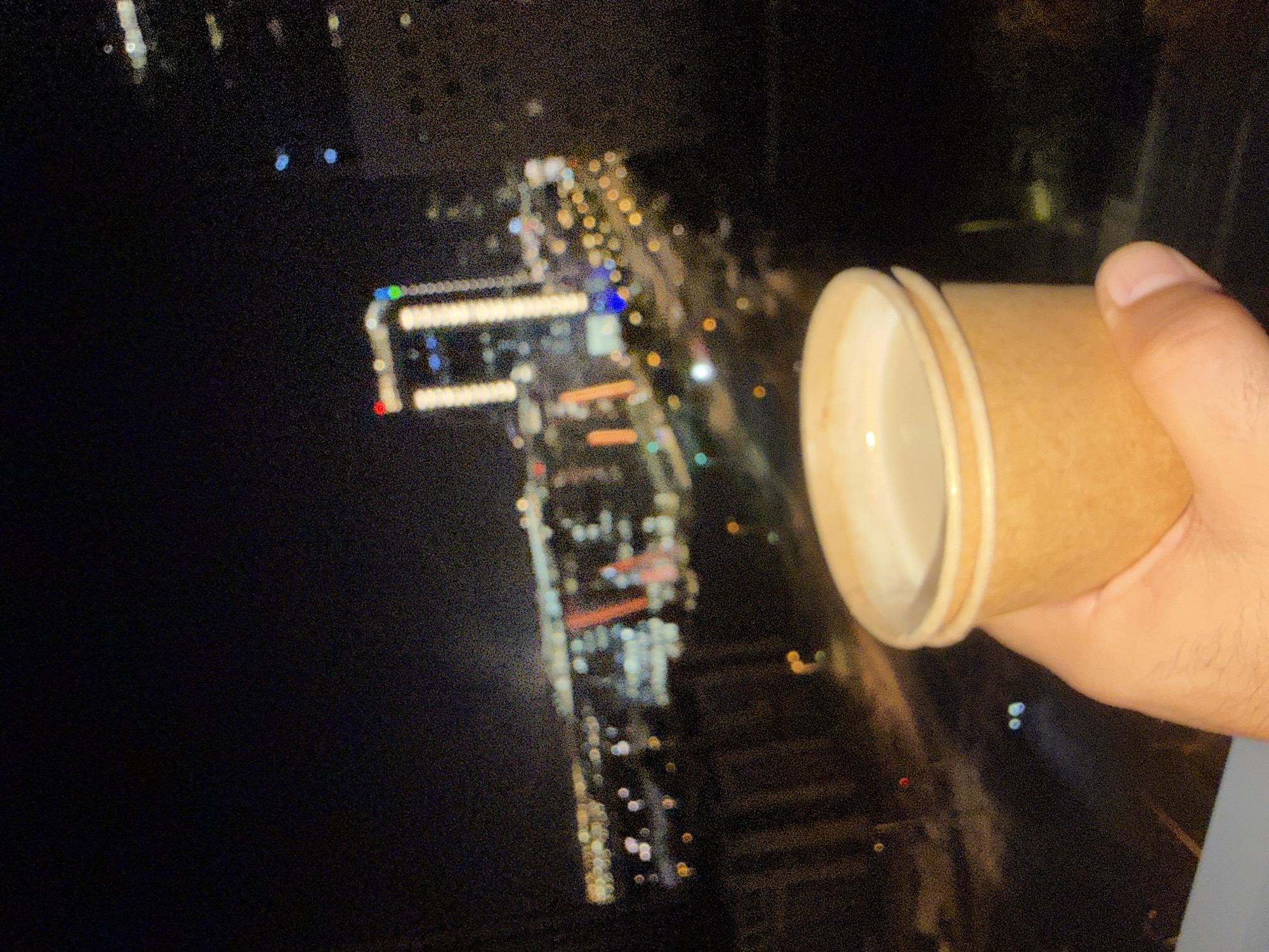 A hand holds a paper cup filled with a light-colored liquid, likely a beverage, against a blurred, nighttime cityscape. The cup is positioned in the foreground, partially obscuring the background. The city lights are visible in the distance, creating a bokeh effect. The image appears to have been taken from a high vantage point, such as a building or balcony.