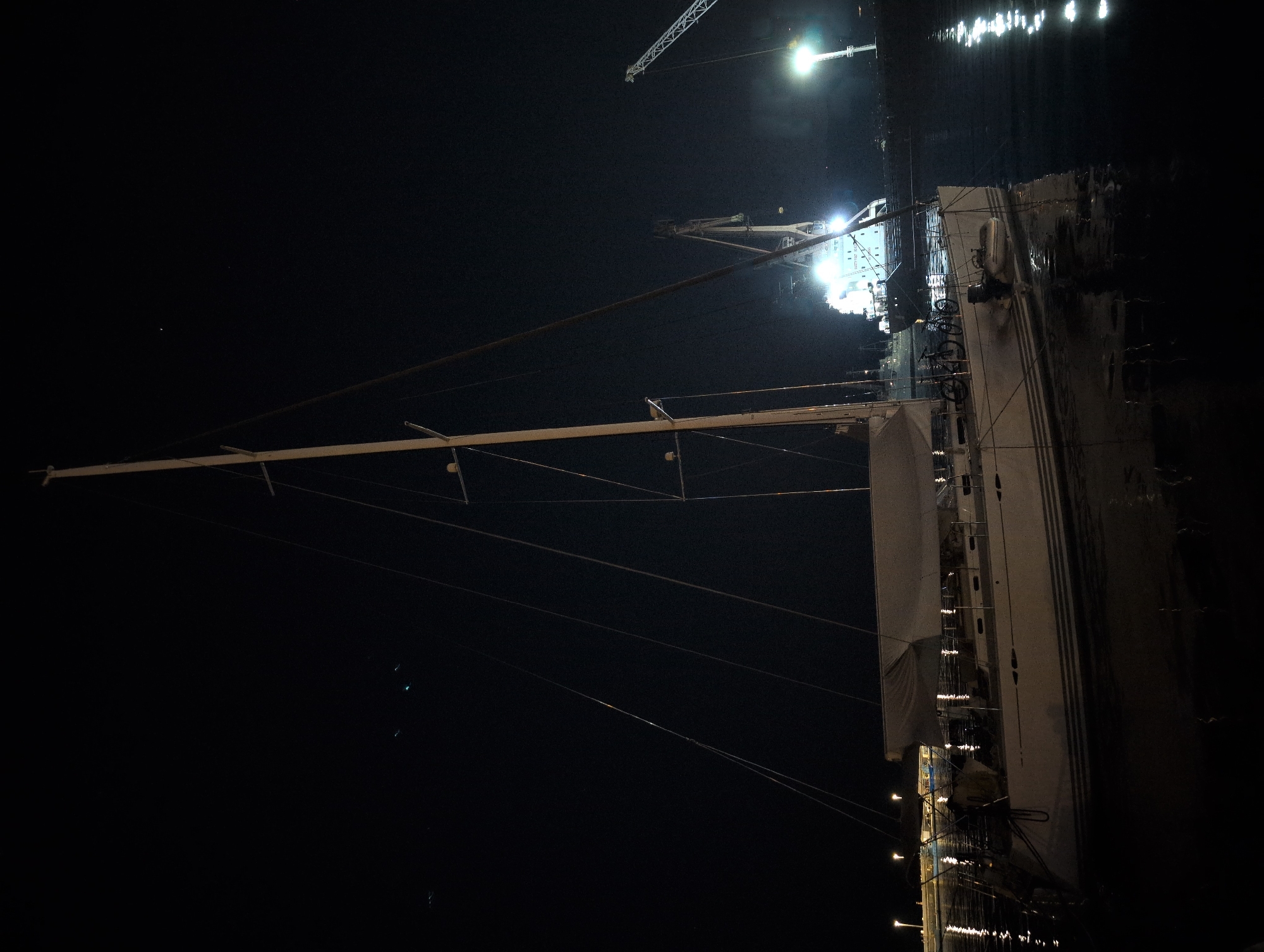 A white sailboat is moored at night in a harbor, possibly in the city of Grad Rijeka, Croatia. The sailboat, which is the primary subject, is positioned in the foreground and takes up a significant portion of the image. It has a tall mast and is covered by a sail. Two bicycles and a small inflatable boat are visible on its deck. The water surrounding the sailboat reflects the lights from the harbor, creating a shimmering effect. In the background, a large cargo ship is illuminated by bright lights, and industrial cranes are visible against the dark sky. The overall scene is dimly lit, with artificial lights casting long reflections on the water.