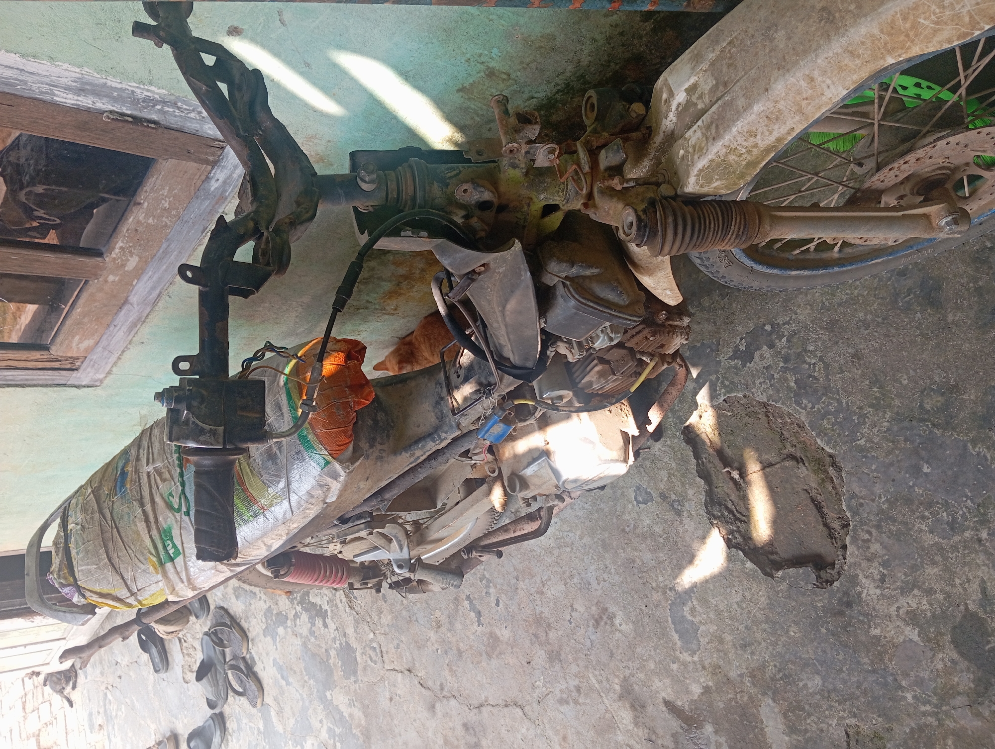 A motorcycle is parked on a concrete ground next to a light green wall. The motorcycle appears old and worn, with some parts missing or damaged. The seat is covered with a woven plastic bag. In the background, there is a wooden window frame with glass panes. There is a gap in the concrete ground to the left of the motorcycle. A ginger cat is visible near the wall behind the motorcycle. Several pairs of sandals are scattered on the ground to the left of the motorcycle. The scene is lit by sunlight, casting shadows on the ground.