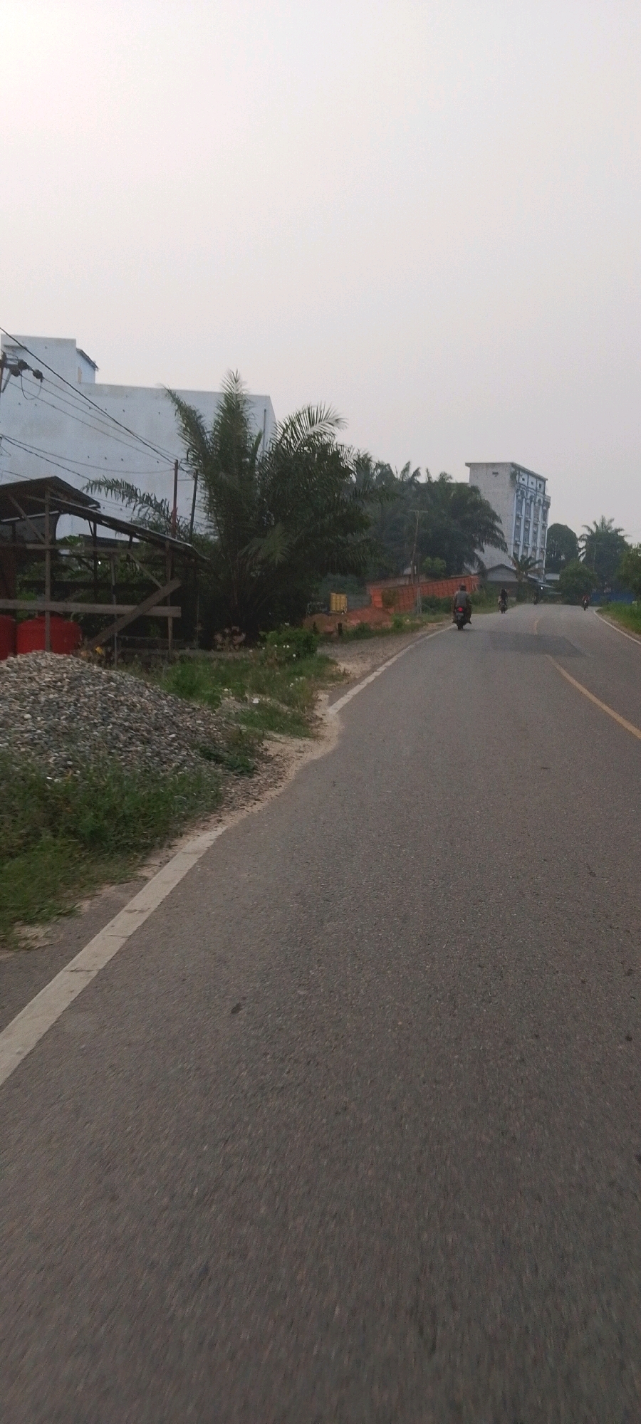 The image captures a roadside scene with a paved road curving to the right. On the left side of the road, there is a pile of grey gravel and a simple wooden structure with a corrugated roof. Beyond this structure, a large, light-colored building with many windows is visible, along with palm trees and other vegetation. In the distance, a taller, white building with blue trim stands out against the hazy sky. Several motorcycles are present on the road, with one rider clearly visible in the foreground. The overall environment suggests an urban or semi-urban setting in Indonesia, possibly Lenggadai Hilir, Indonesia, characterized by a mix of construction materials, vegetation, and buildings alongside a public thoroughfare.