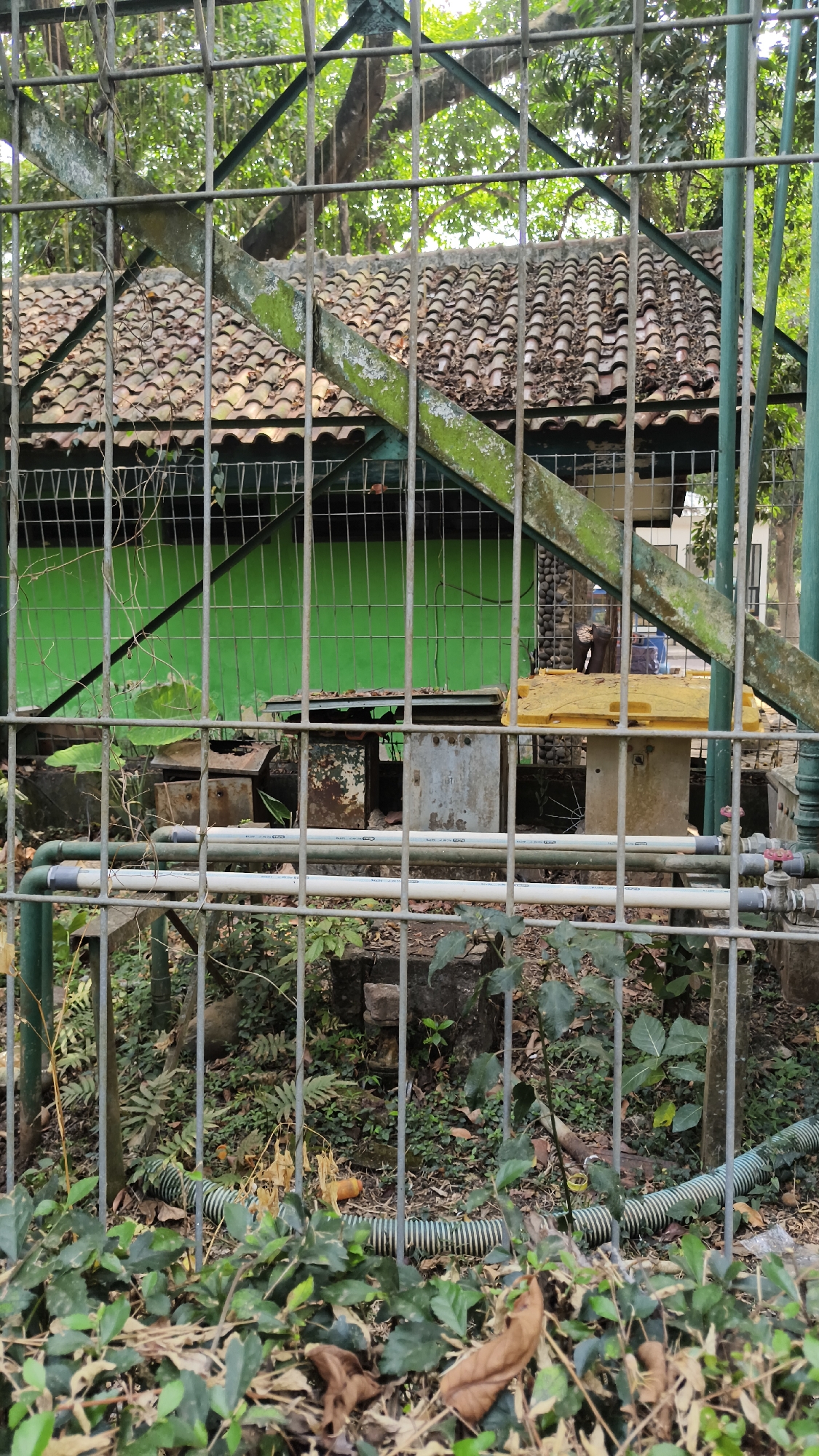The image depicts an outdoor scene behind a metal fence, likely in South Jakarta, Indonesia. In the background, a building with a tiled roof is visible, painted bright green. Some weathered metal structures and a yellow container are present in front of the building. Two horizontal pipes run across the foreground, supported by the fence. Green foliage and some dead leaves are scattered on the ground. The overall impression is of a slightly neglected or storage area within an urban environment.
